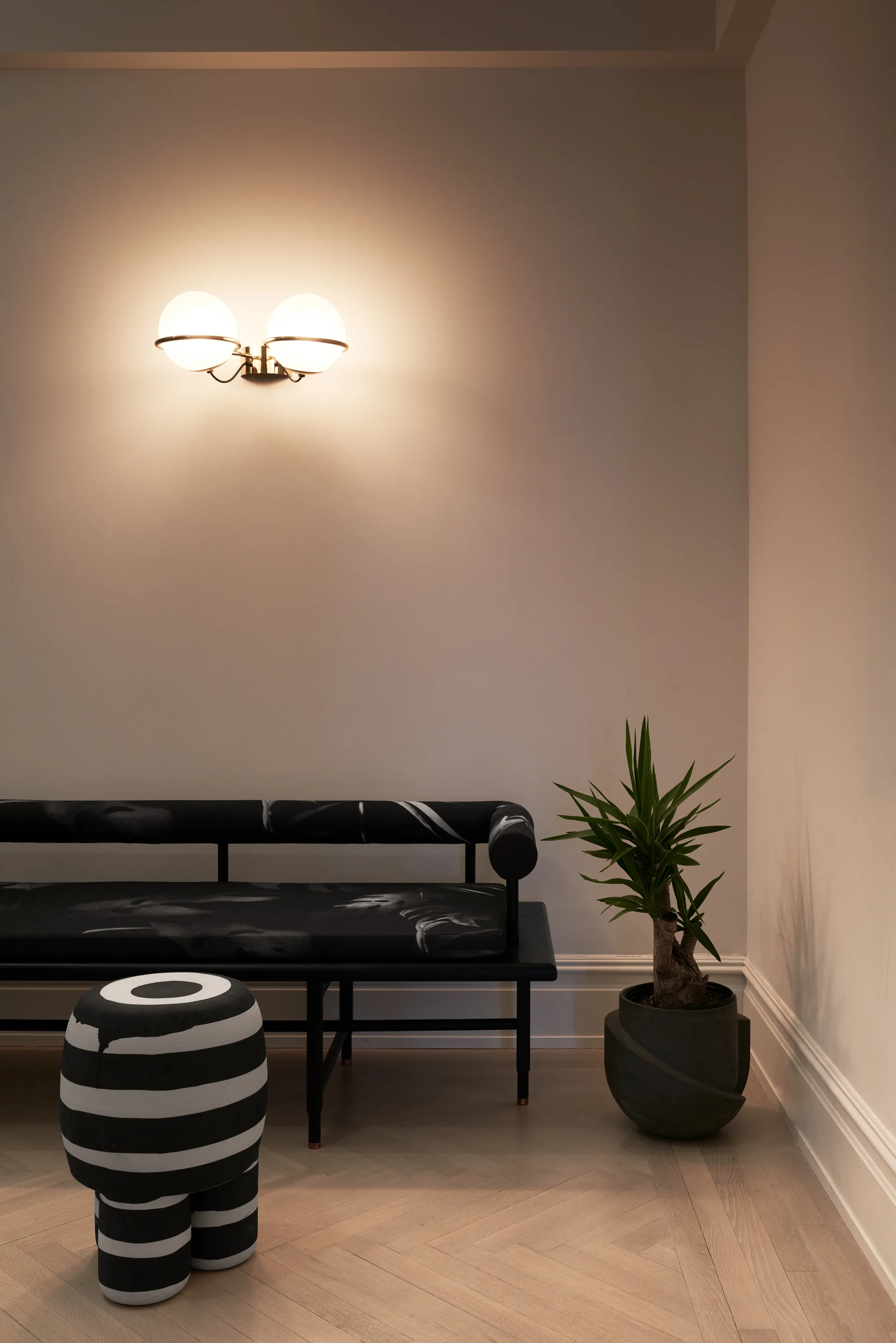 A minimalist corner features a black patterned daybed, a black and white striped stool, a potted plant, and a glowing globe sconce on the wall.