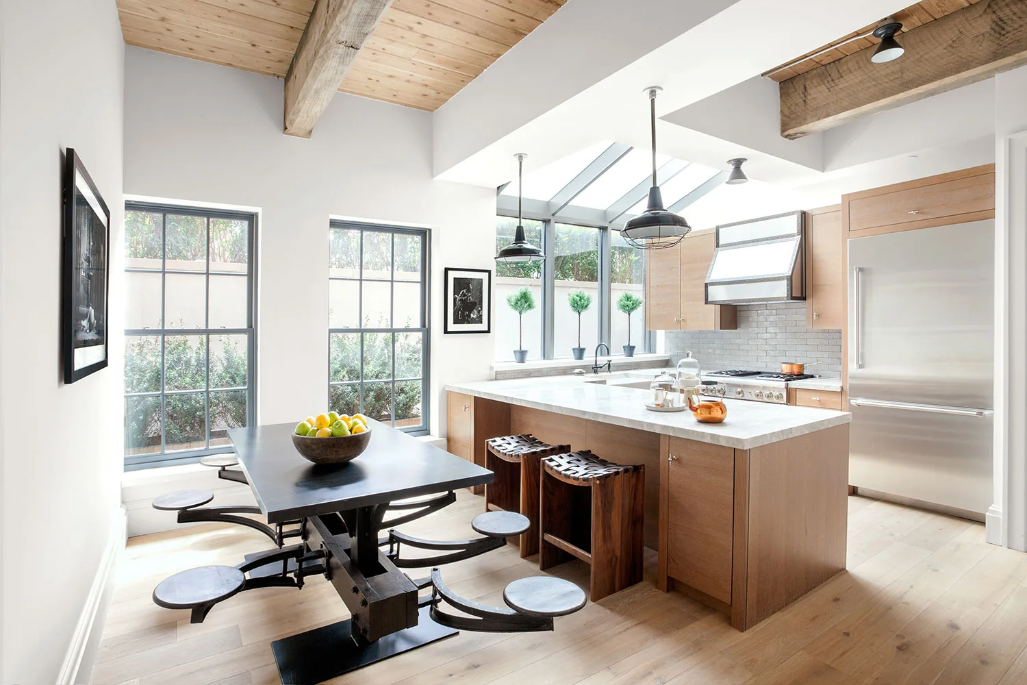 A bright kitchen features a light wood island, a black industrial-style dining table with attached swing-out stools, and exposed wood ceiling beams.