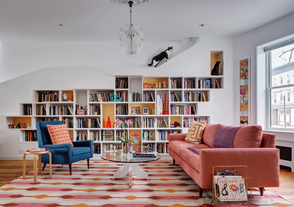 An eclectic living room with a white stepped built-in bookcase, a coral pink sofa, a blue armchair, and a colorful geometric patterned rug.