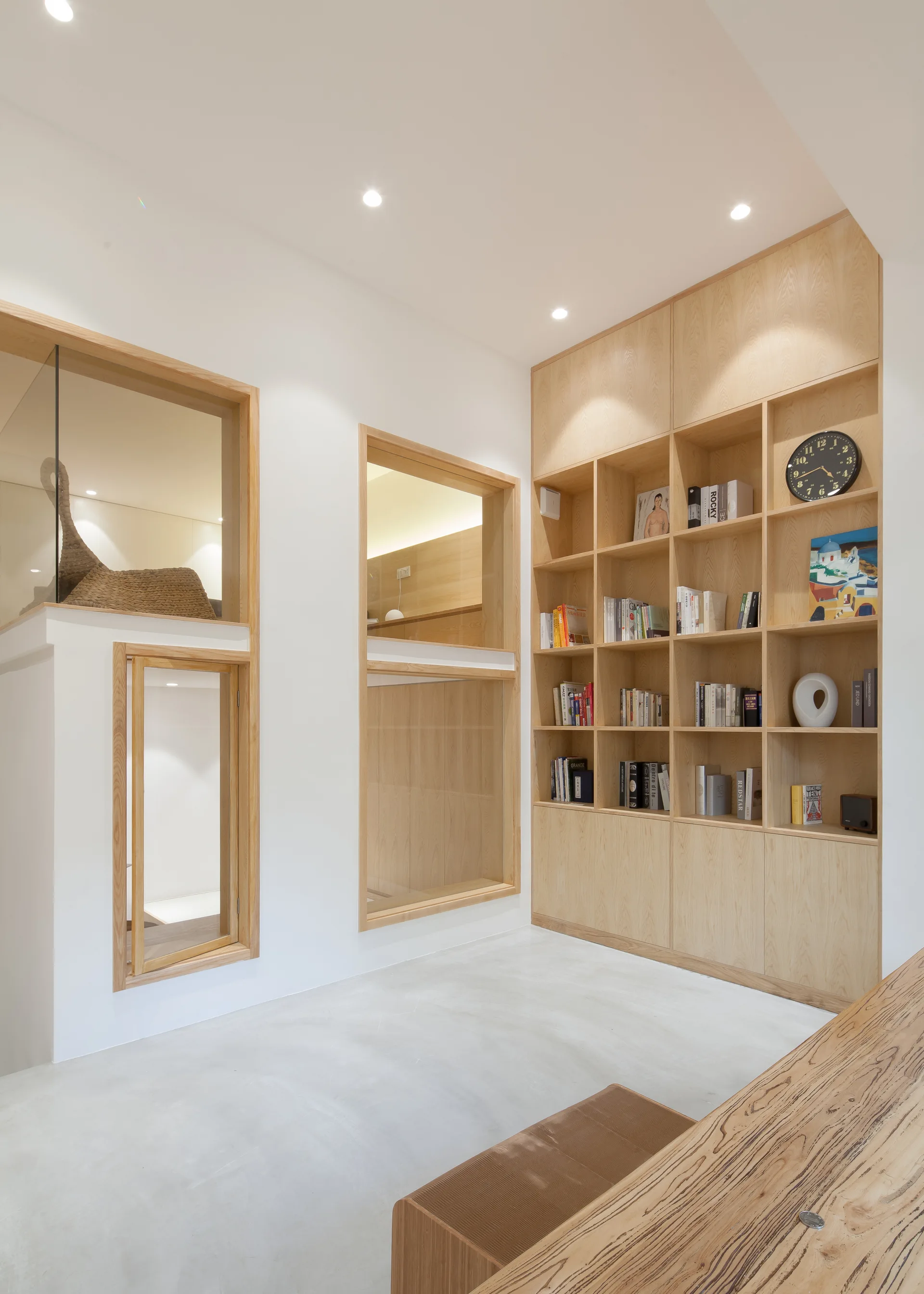 A minimalist interior features a full-height light wood bookshelf, clean white walls, and interior windows framed in matching wood.