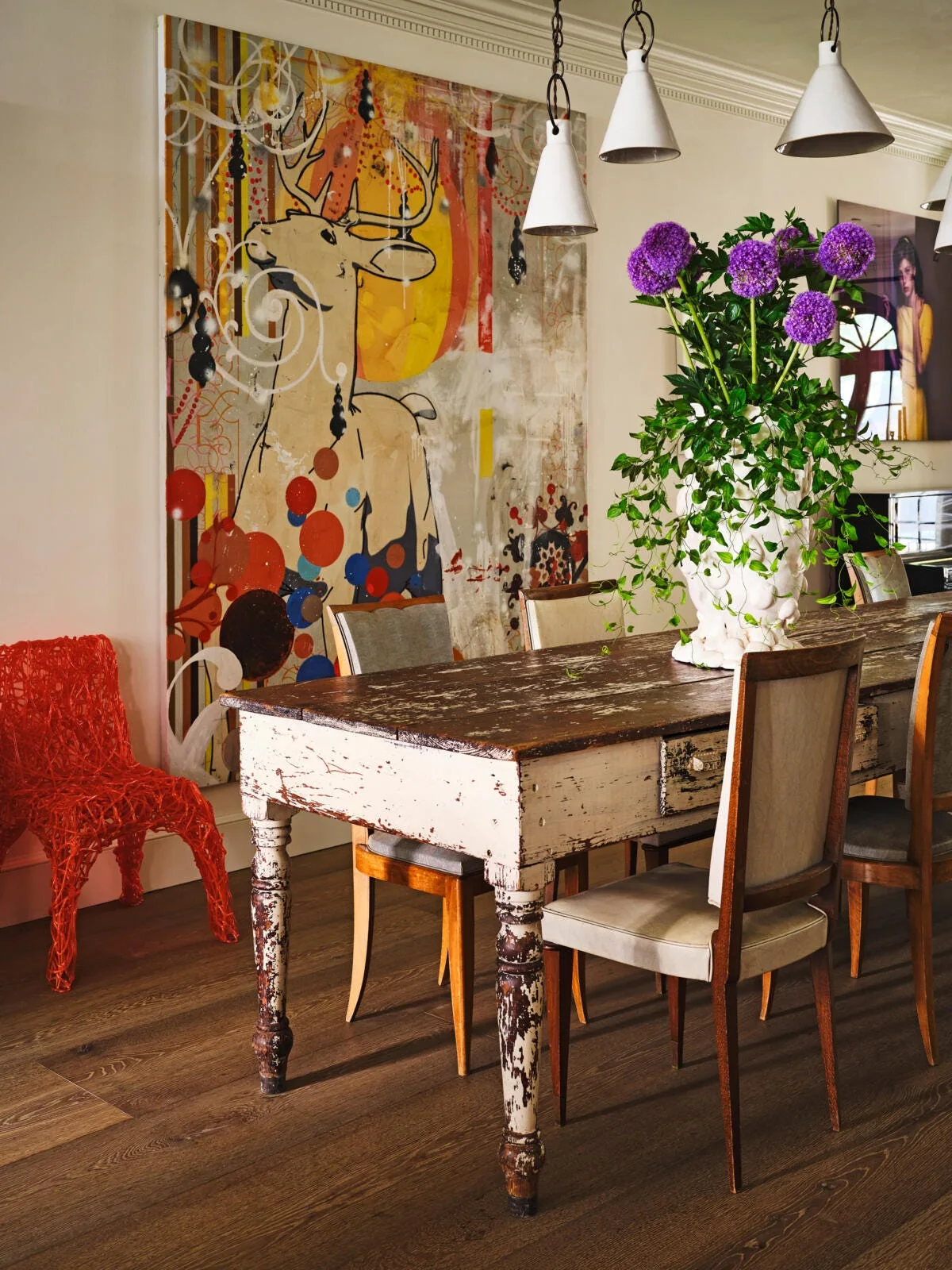 An eclectic dining room featuring a distressed wooden table, upholstered chairs, white cone pendant lights, a large deer painting, and a red textured chair.