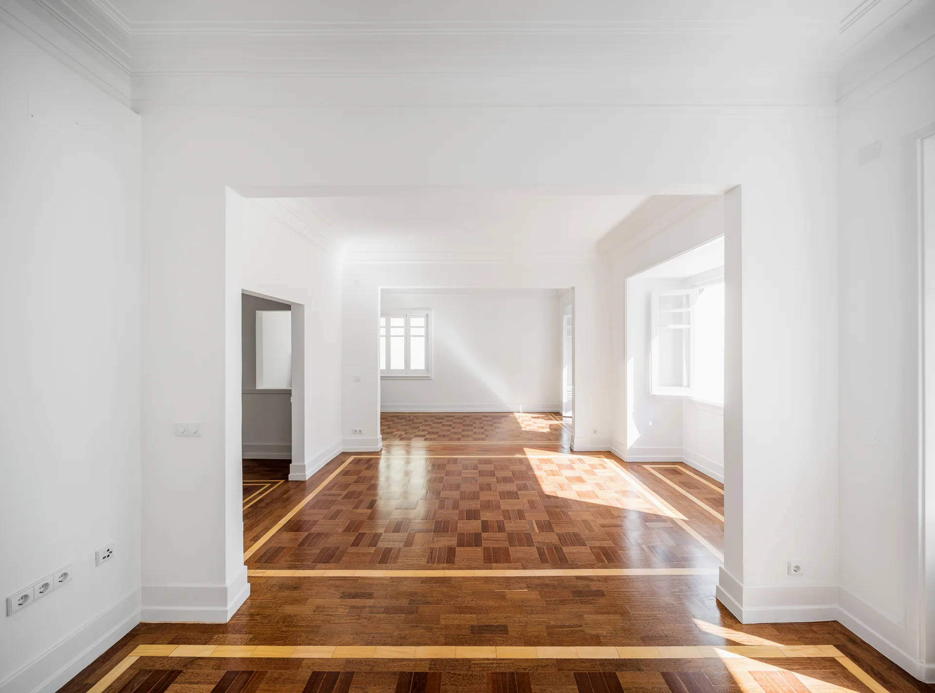 An empty residential room with clean white walls, wide square archways, and patterned brown parquet wood flooring with light borders.
