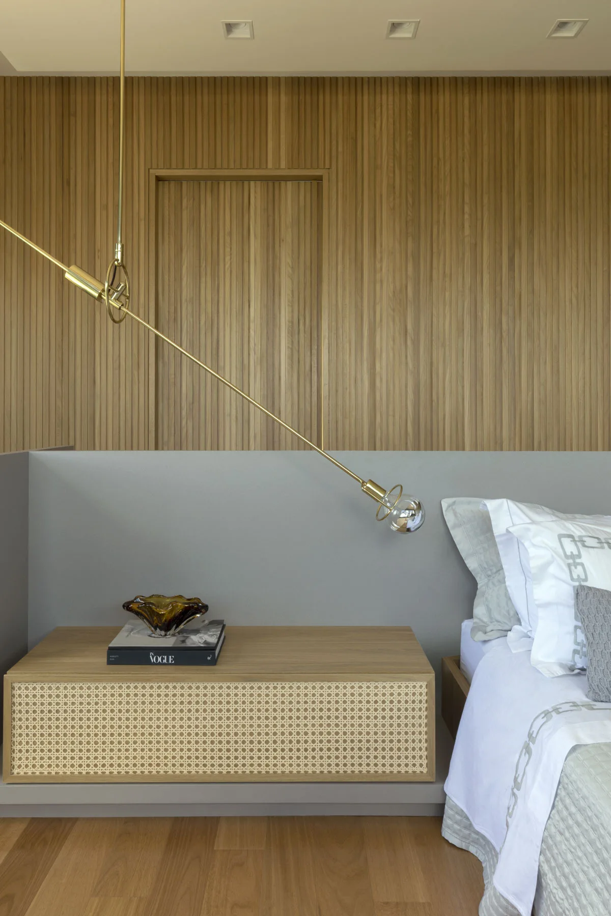 A modern bedroom features a vertical slatted wood accent wall, a low grey headboard, a wooden nightstand with a cane front, and an angled brass light.