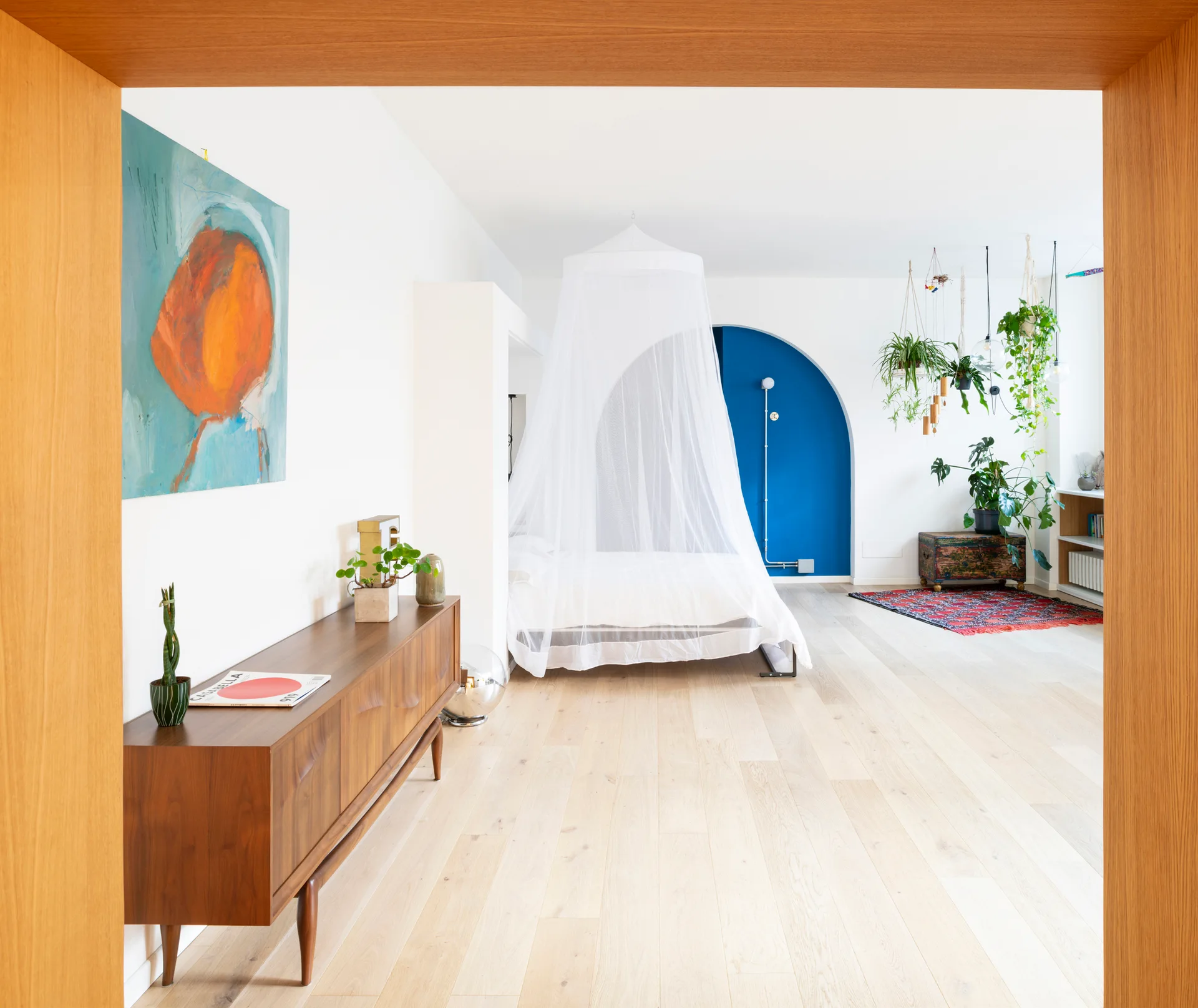An airy bedroom framed by a wooden doorway, featuring a mid-century credenza, a canopy bed, and a cobalt blue arched wall niche.