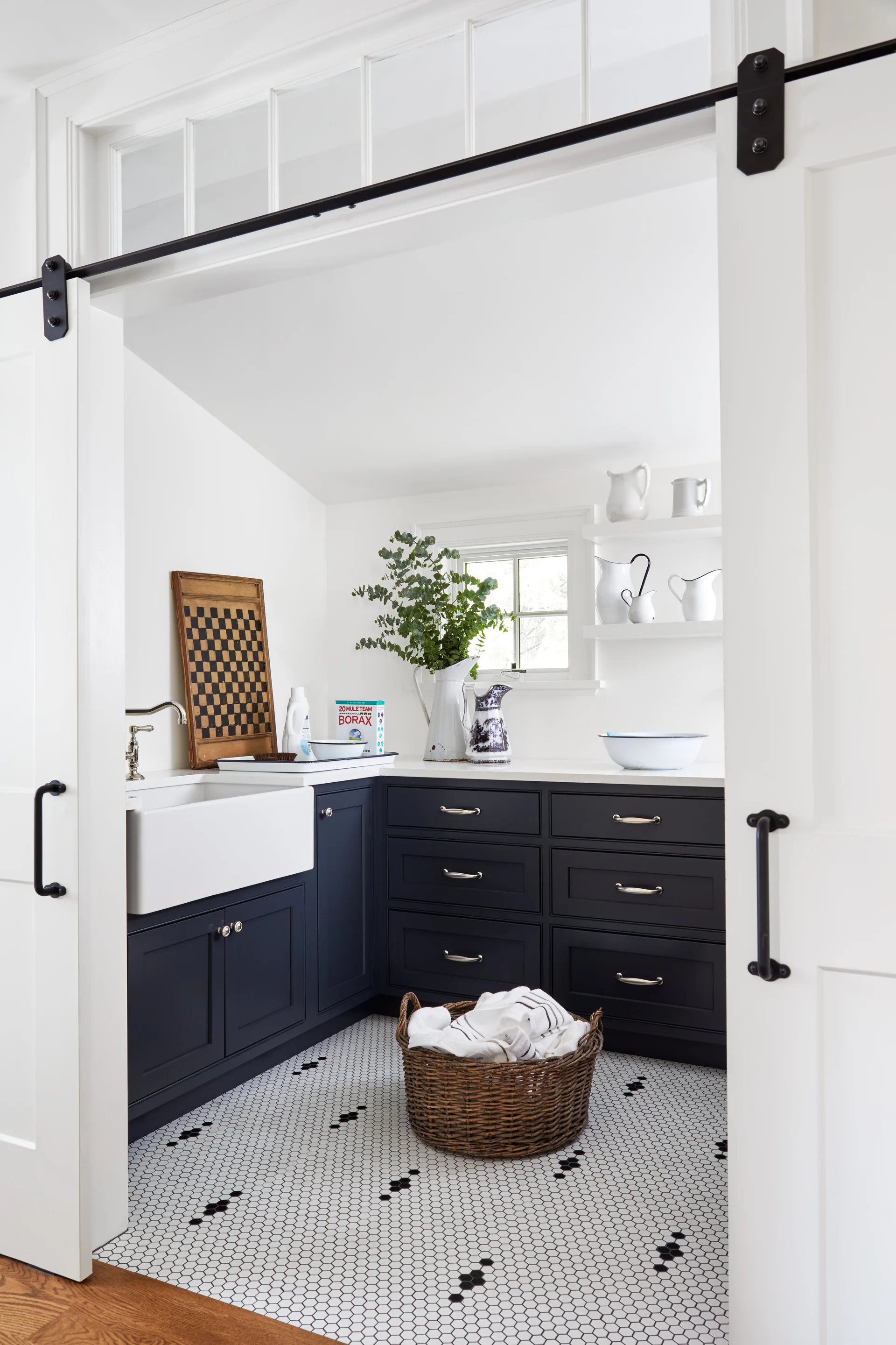 A modern farmhouse laundry room with navy blue cabinetry, a white apron sink, patterned hexagonal tile floors, and open sliding white barn doors.