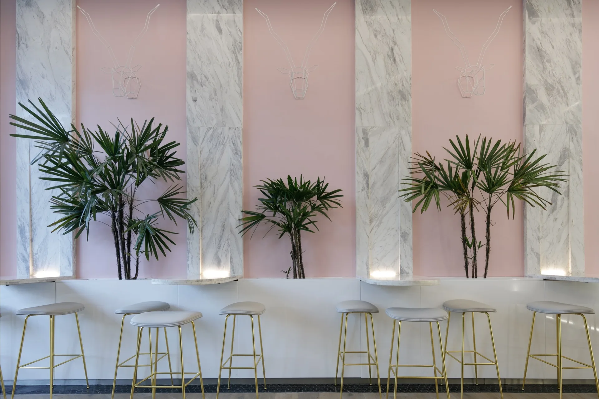 A modern bar seating area featuring a row of gold-legged stools against a white counter under alternating pink and white marble wall panels.