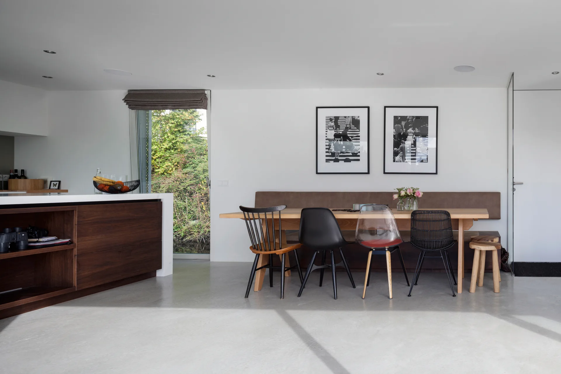 A modern dining room features a wooden table, an upholstered wall bench, and a collection of mismatched chairs on a smooth concrete floor.