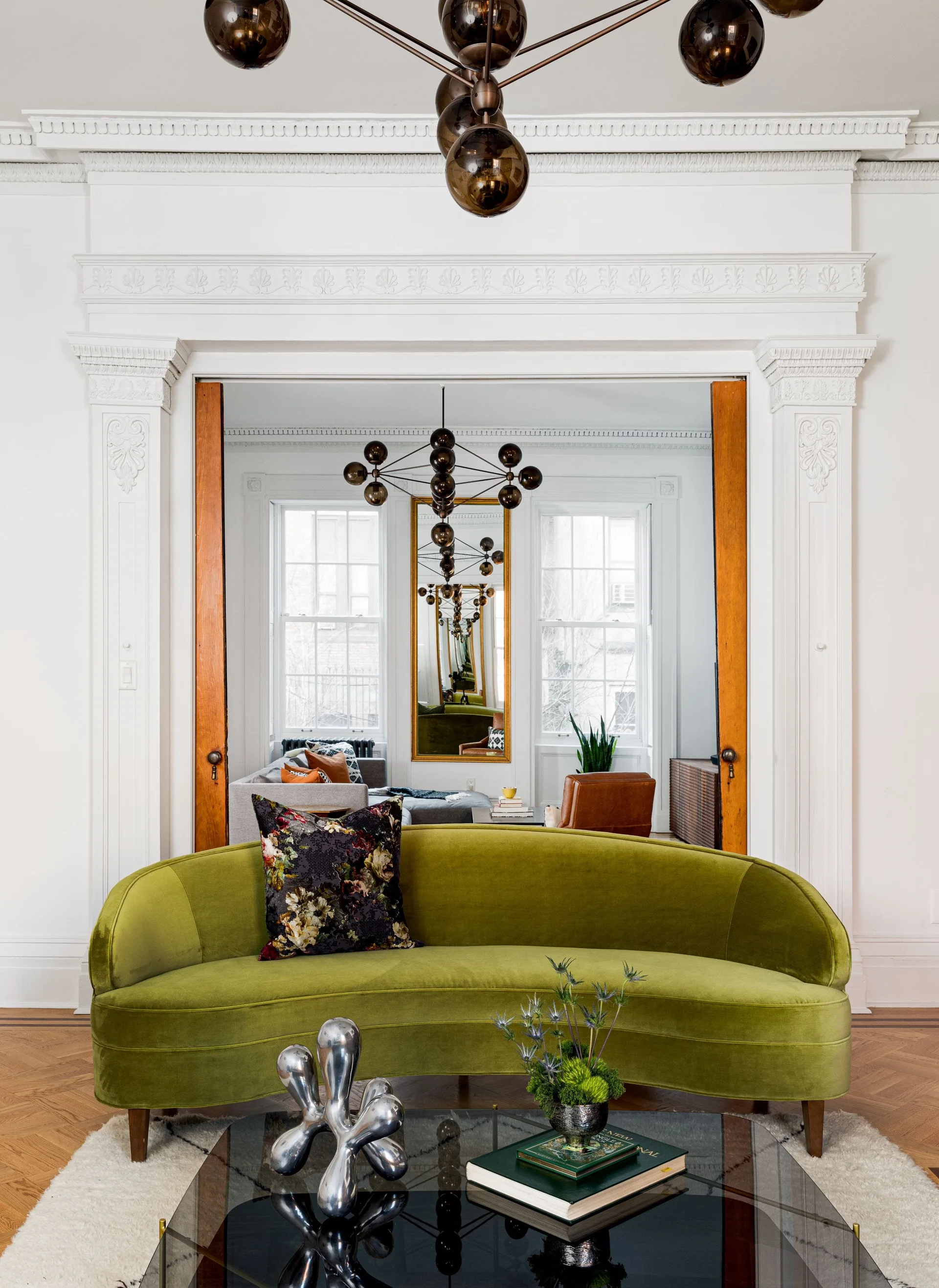 An ornate white architectural doorway with wooden pocket doors frames a curved green velvet sofa and a view into an adjacent seating area.