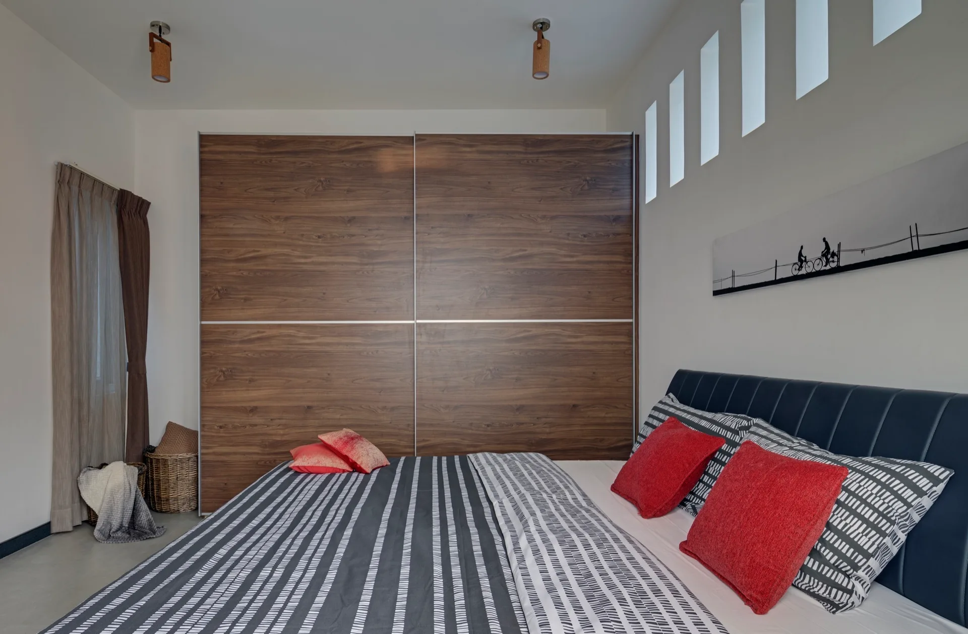 A modern bedroom featuring a large wood-paneled sliding wardrobe, a bed with striped bedding and red cushions, and vertical window slits.