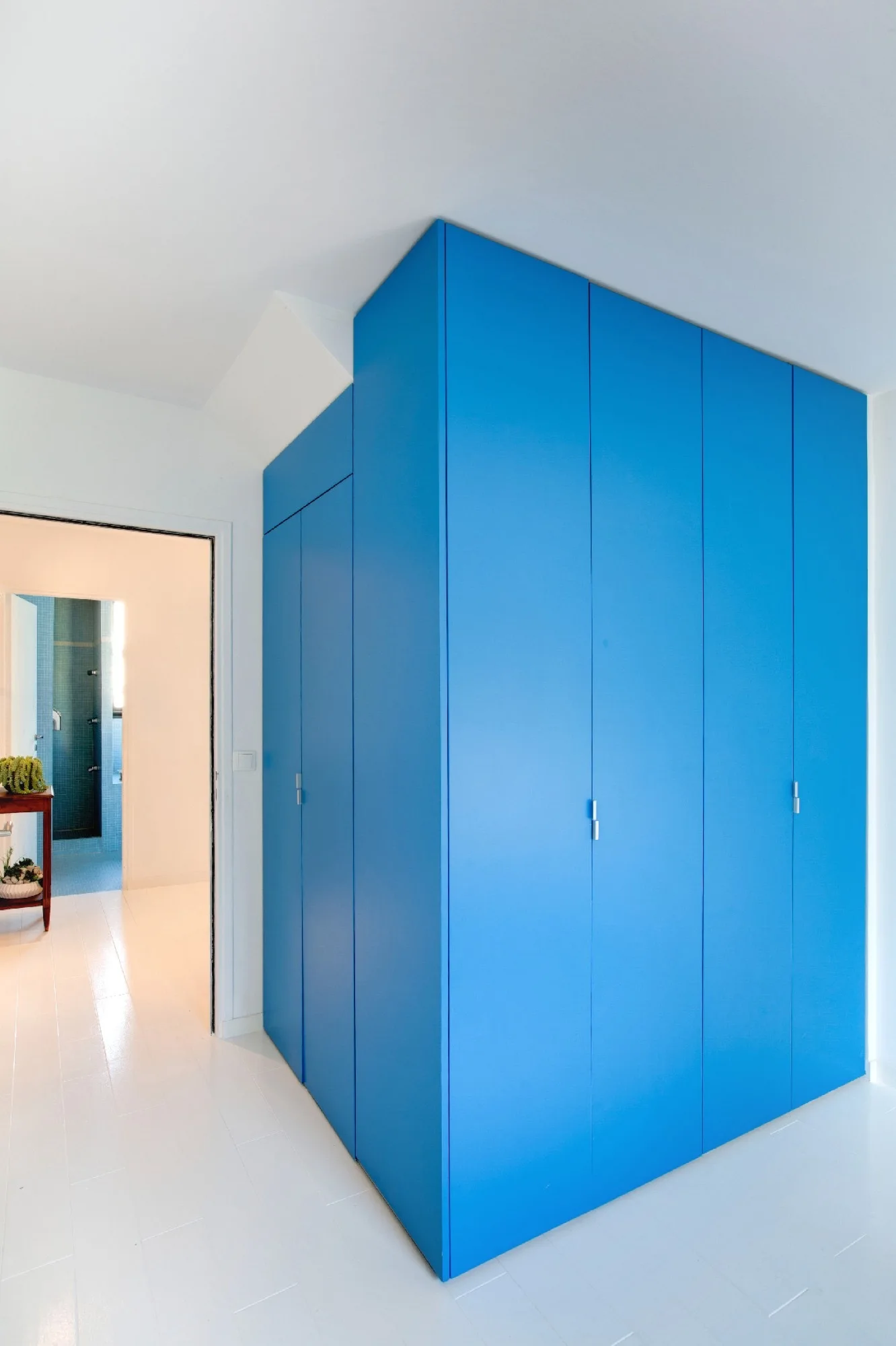 A large floor to ceiling bright blue corner wardrobe with flat panel doors stands on white painted wood floors next to a doorway.