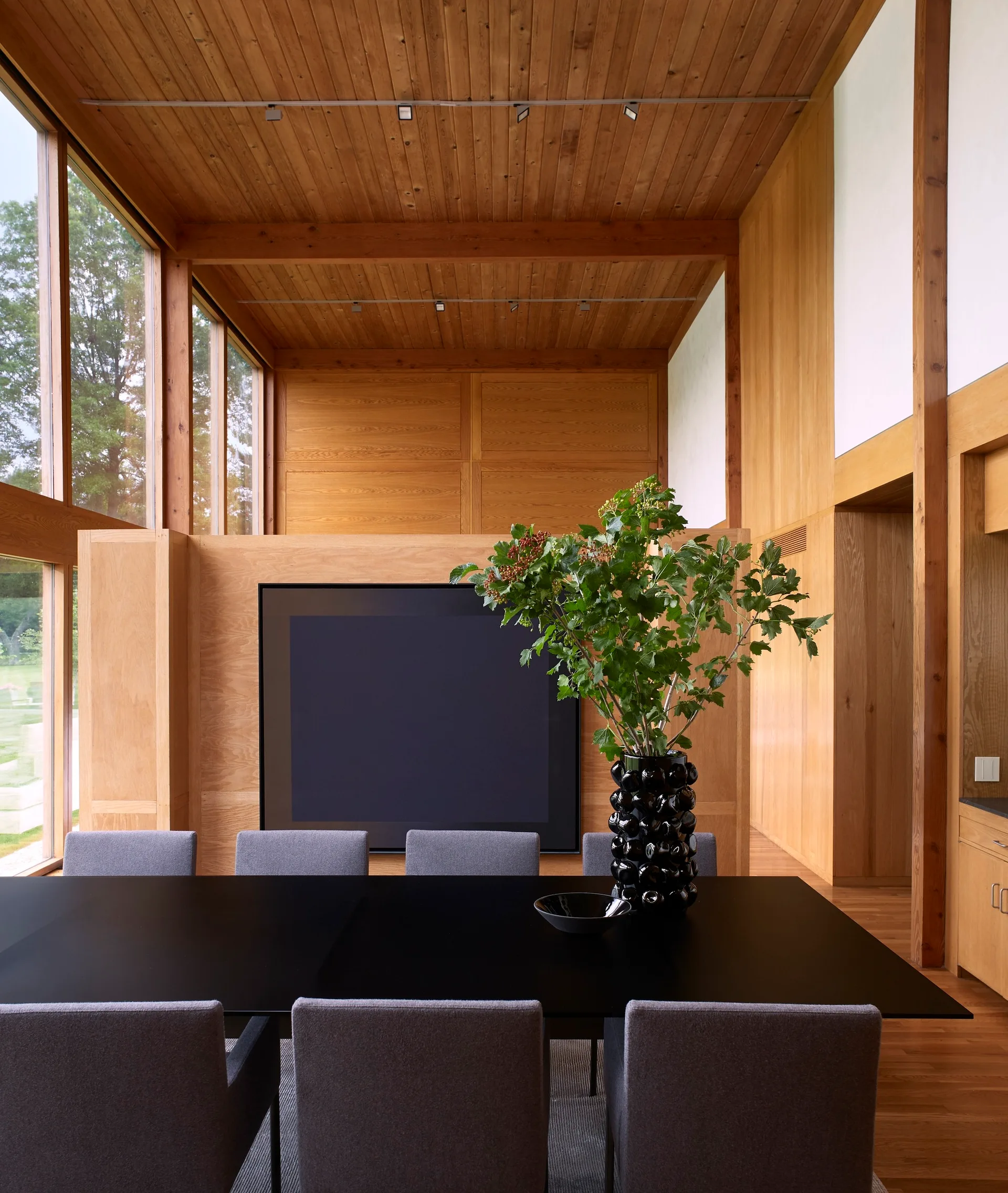 A modern dining room featuring a black table, grey upholstered chairs, and extensive warm wood paneling on the walls and ceiling.