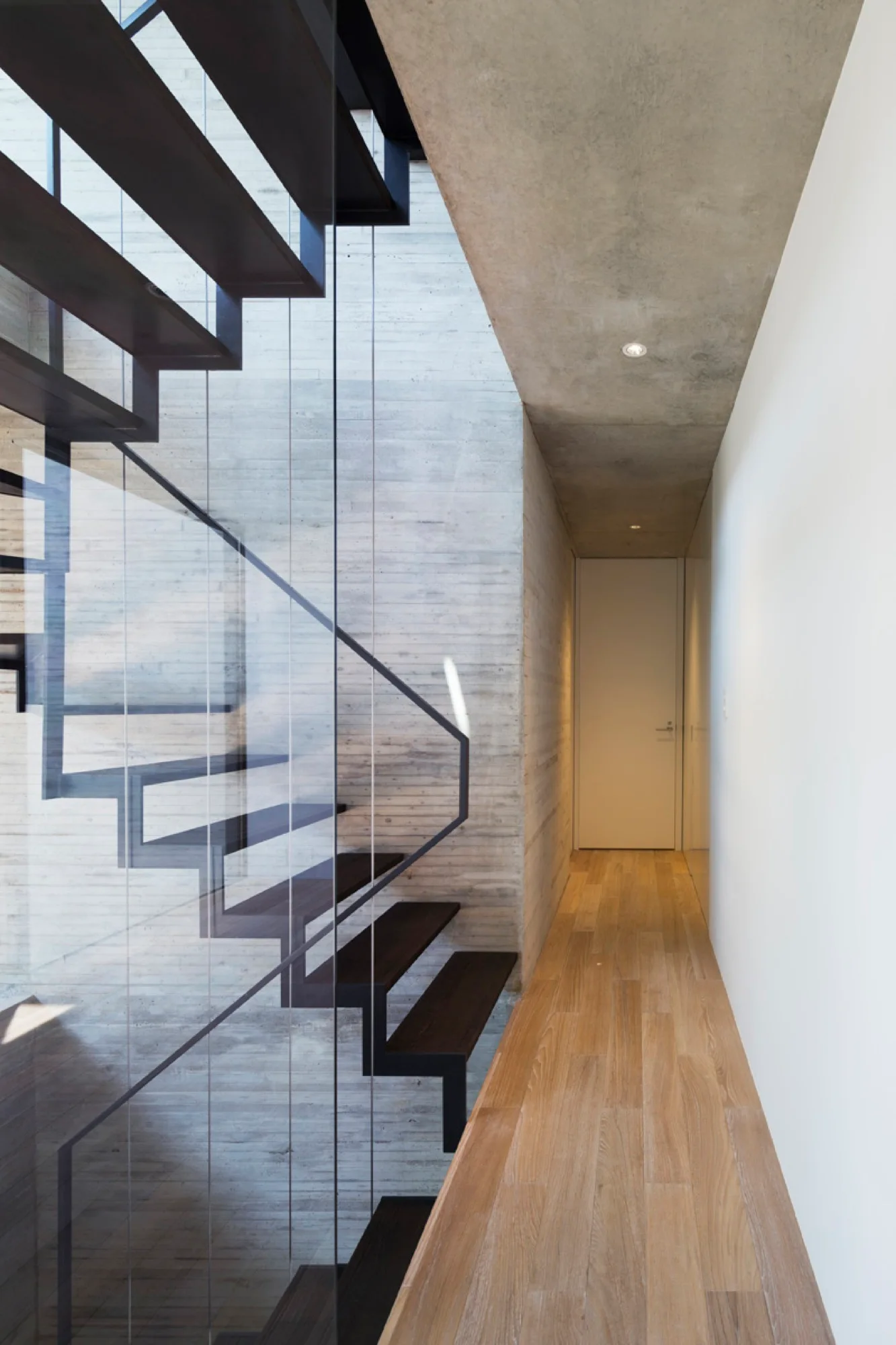 A minimalist hallway features an open riser staircase with dark wood treads and a glass railing next to a board-formed concrete wall.