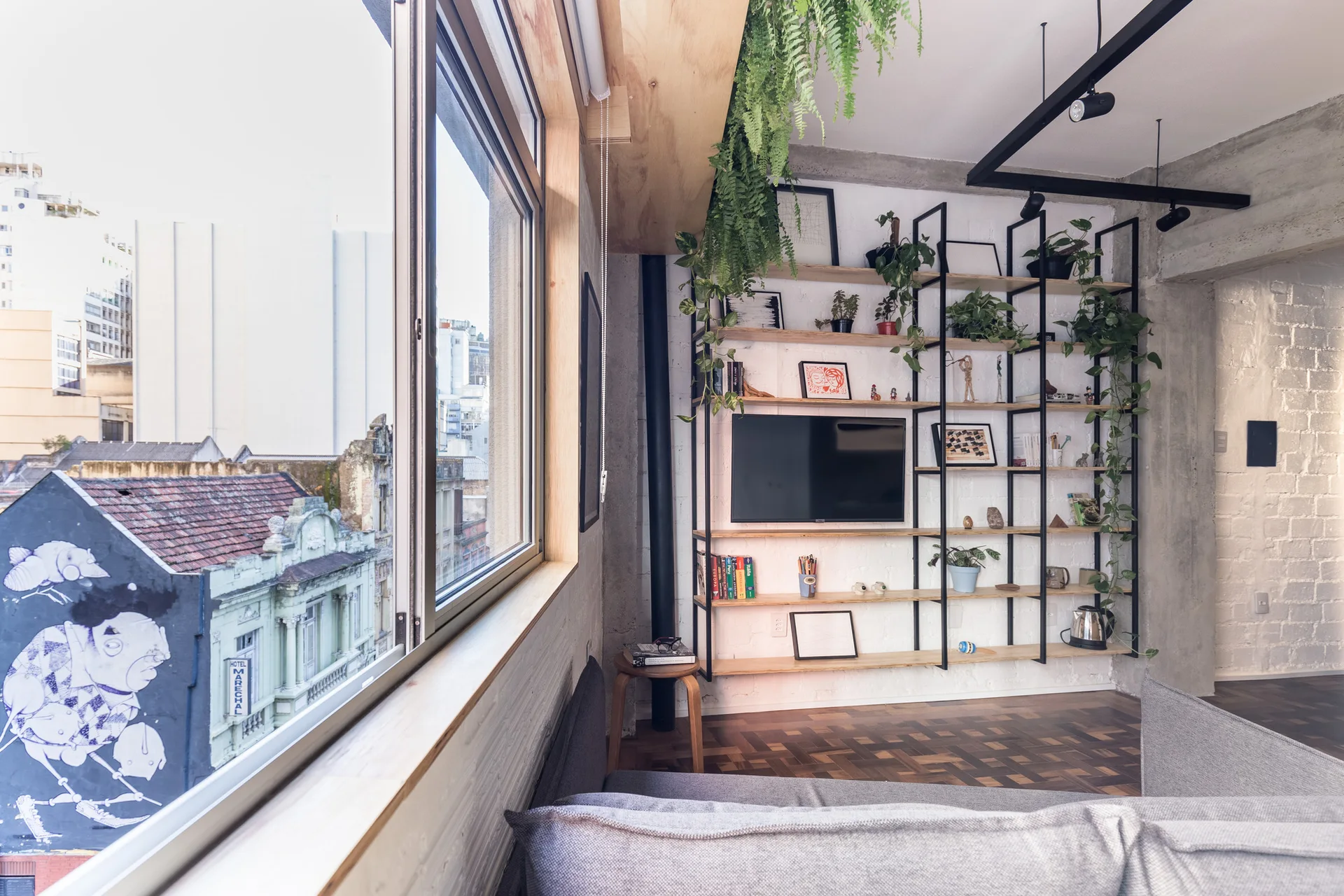 An urban living room features a large window with a city view, a grey sofa, and an industrial metal and wood shelving unit with houseplants.