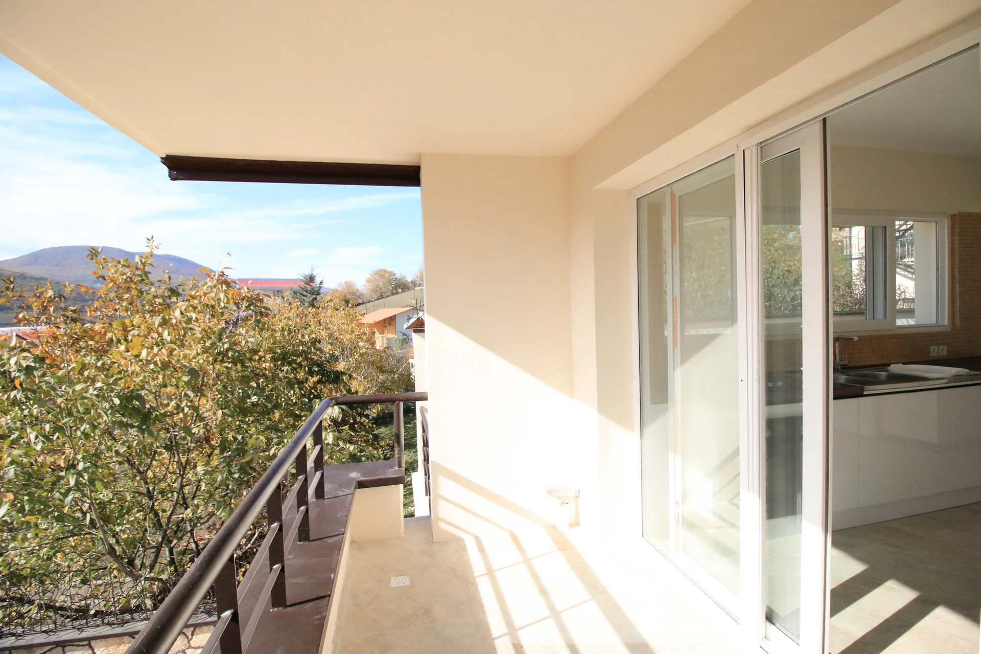 A sunny balcony features beige floor tiles, a dark metal railing, and large sliding glass doors opening to an indoor kitchen.