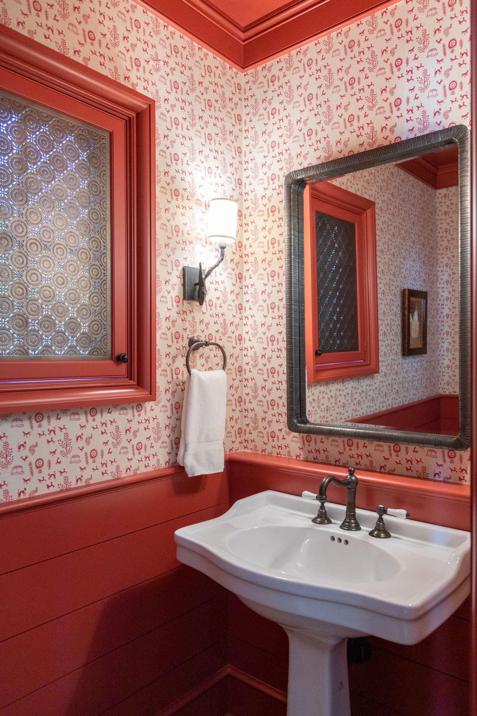 A small powder room features red painted wood wainscoting, whimsical patterned wallpaper, a white pedestal sink, and a dark-framed rectangular mirror.