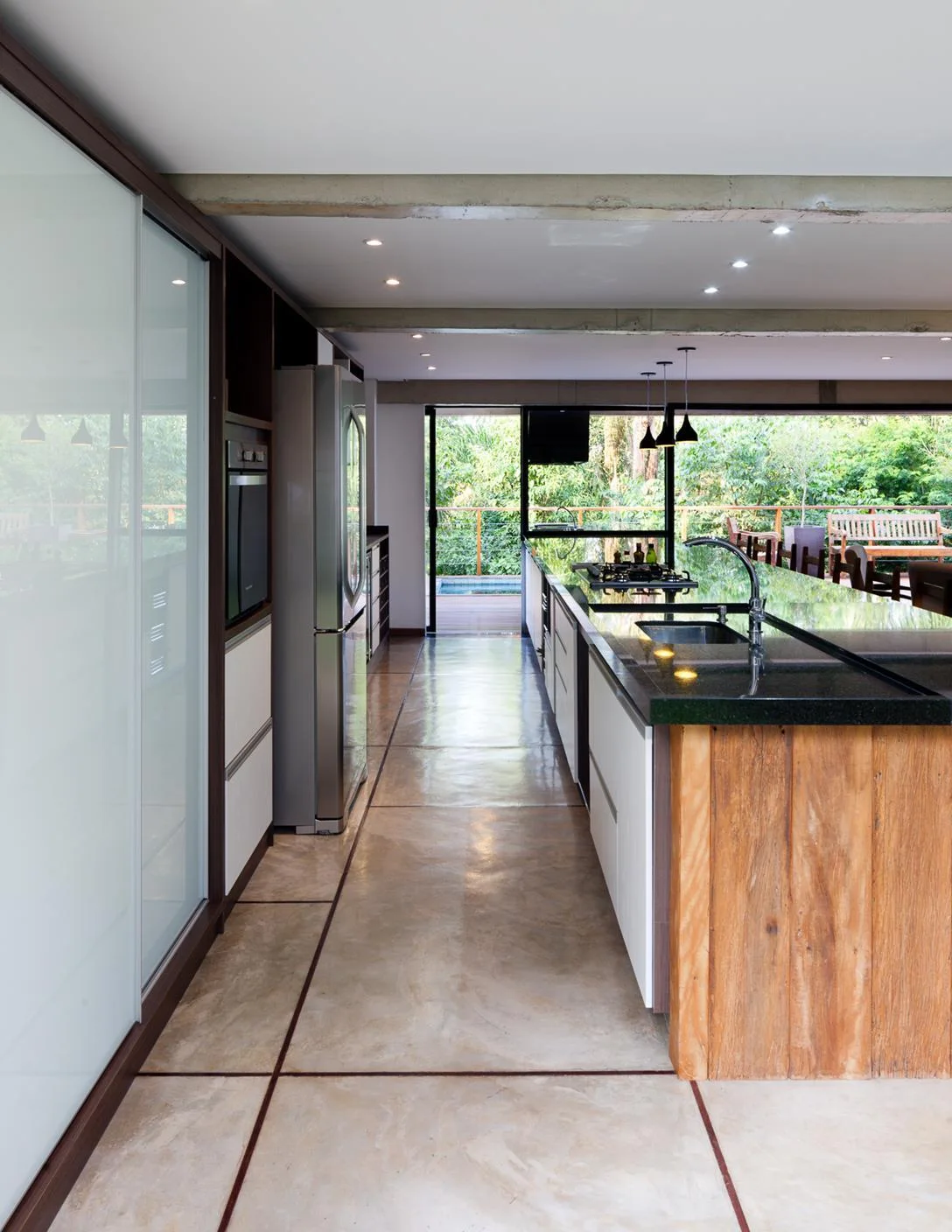 A modern kitchen featuring a long island with a natural wood base, polished concrete tile flooring, and glass doors opening to an outdoor deck.