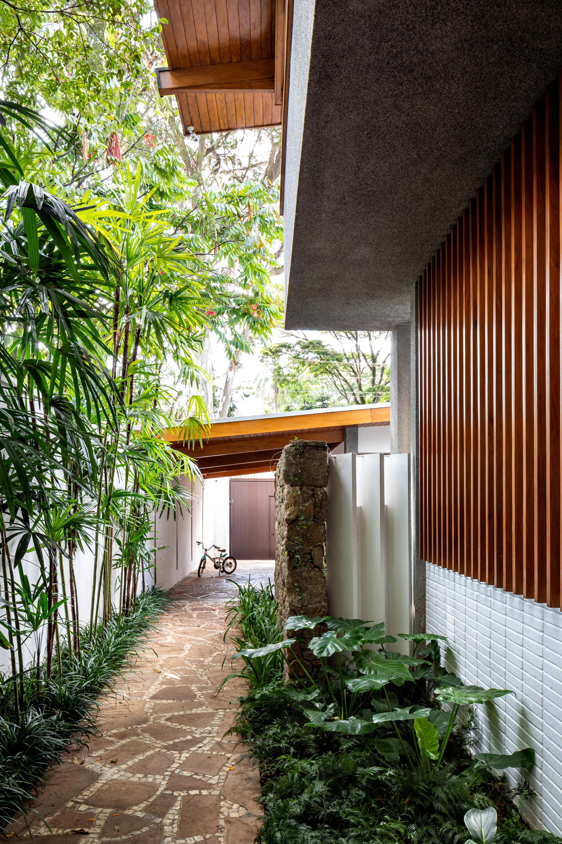 A stone-paved pathway leads through a tropical garden past a modern exterior featuring vertical wood slats and a rustic stone pillar.