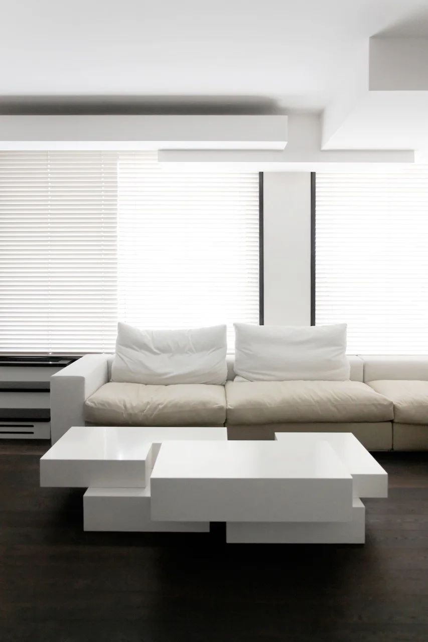 A white modular sofa and a tiered geometric coffee table are set against dark wood flooring and windows with white venetian blinds.