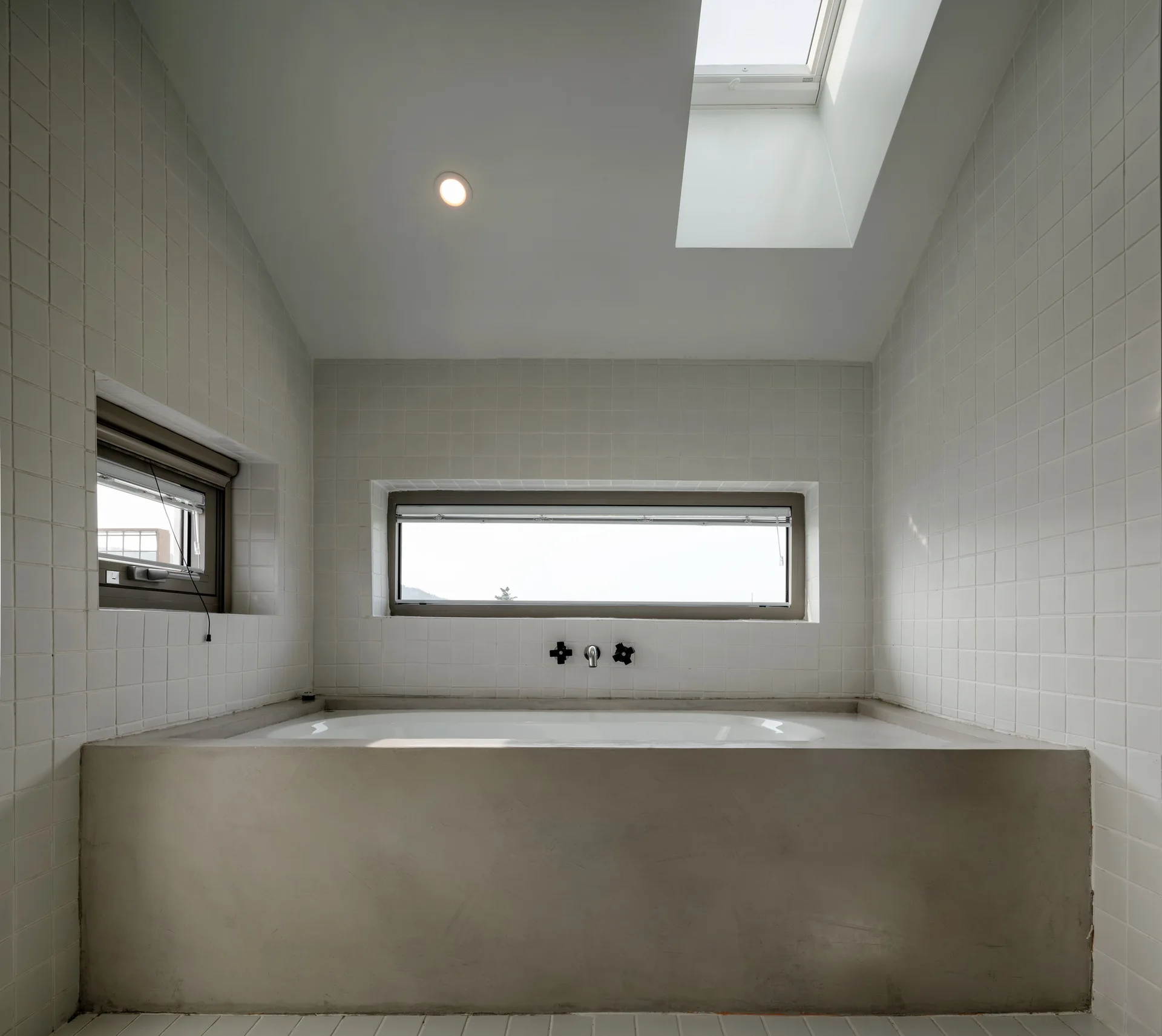 A built-in white bathtub with a smooth grey concrete surround sits beneath a horizontal window in a room with white square grid wall tiles.