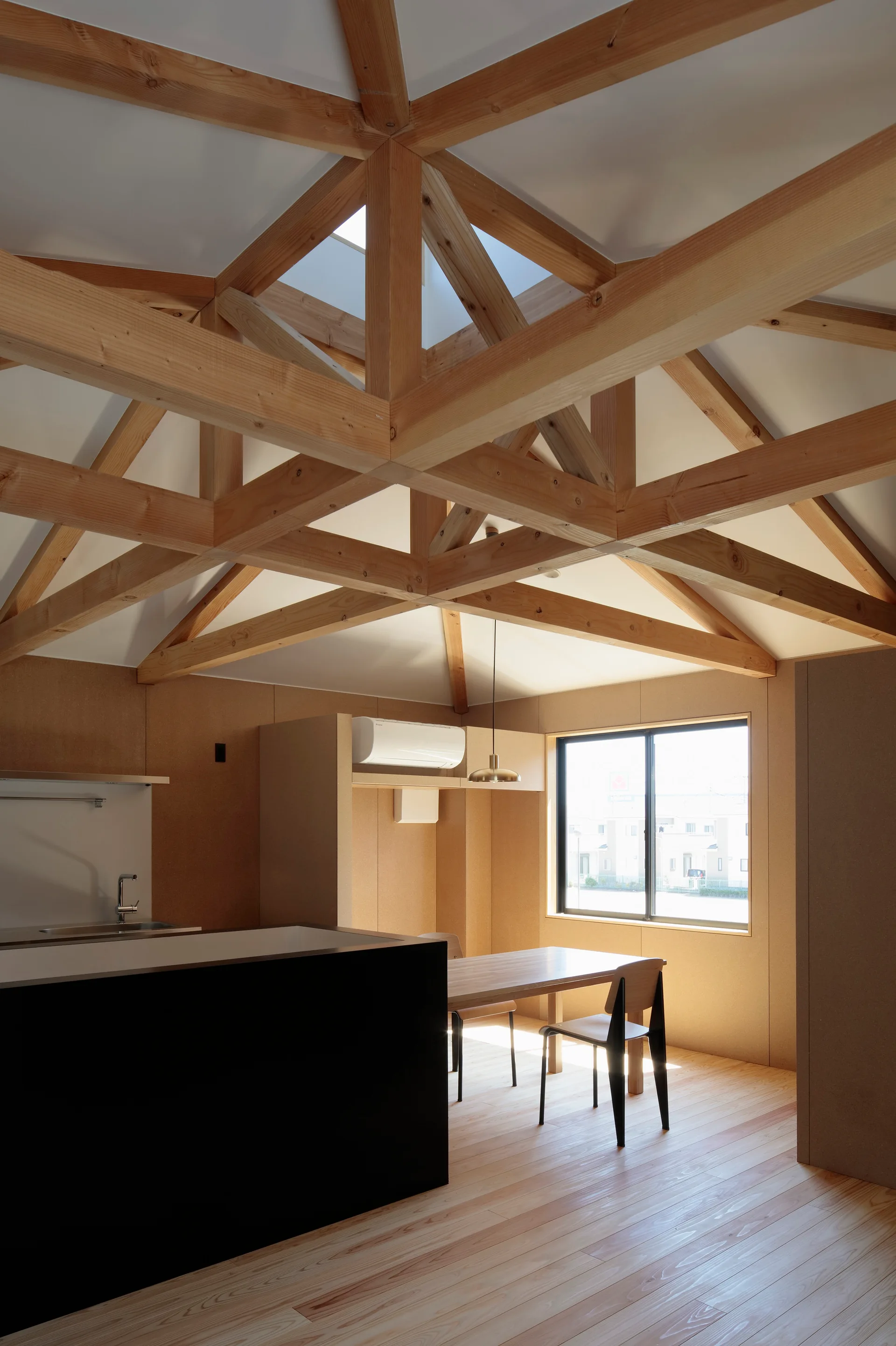 A minimalist dining area and kitchen features an exposed light wood truss ceiling, a black island, and a wooden table by a window.