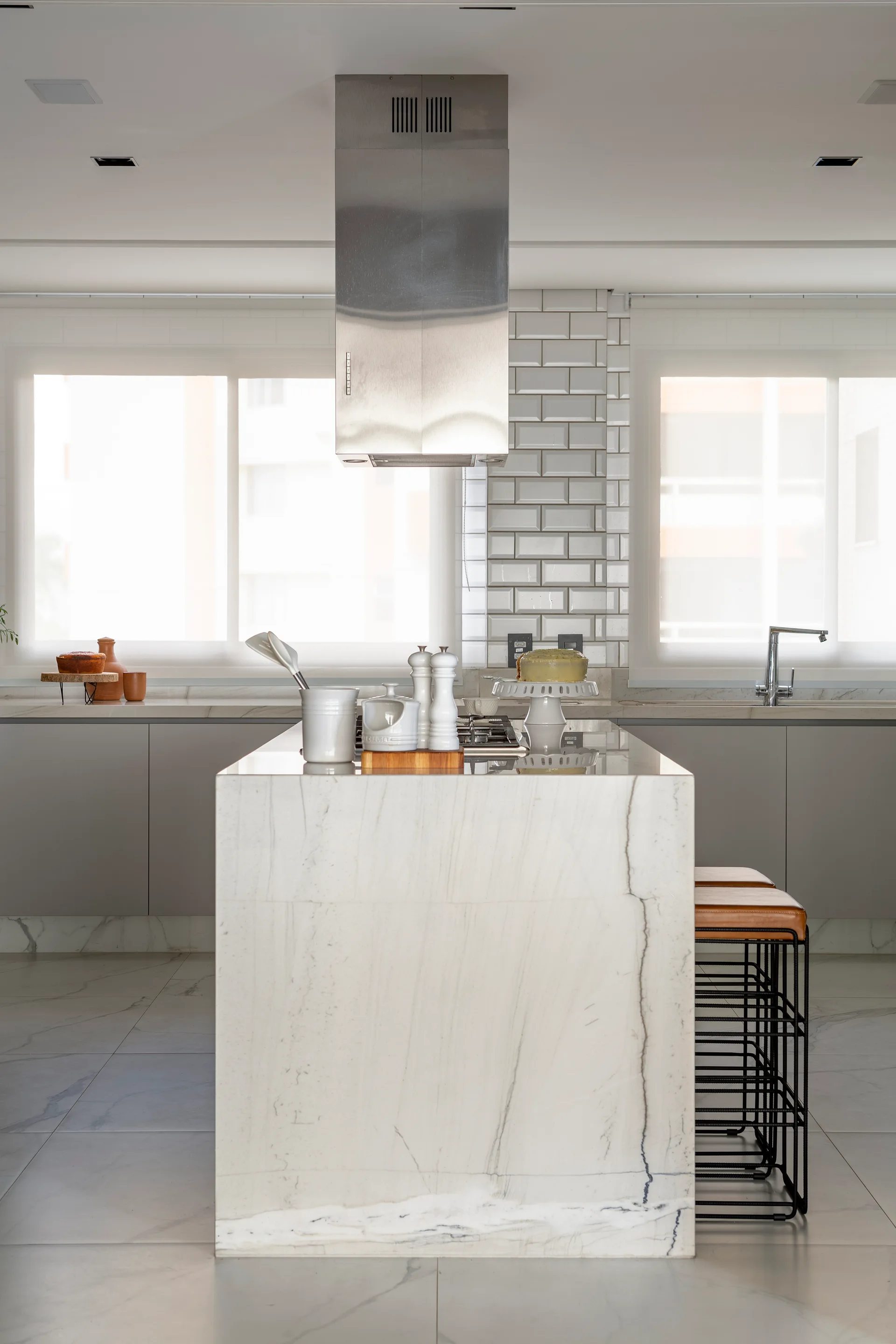 A modern kitchen features a central marble-topped island with a built-in cooktop, a stainless steel range hood, and a white subway tile backsplash.