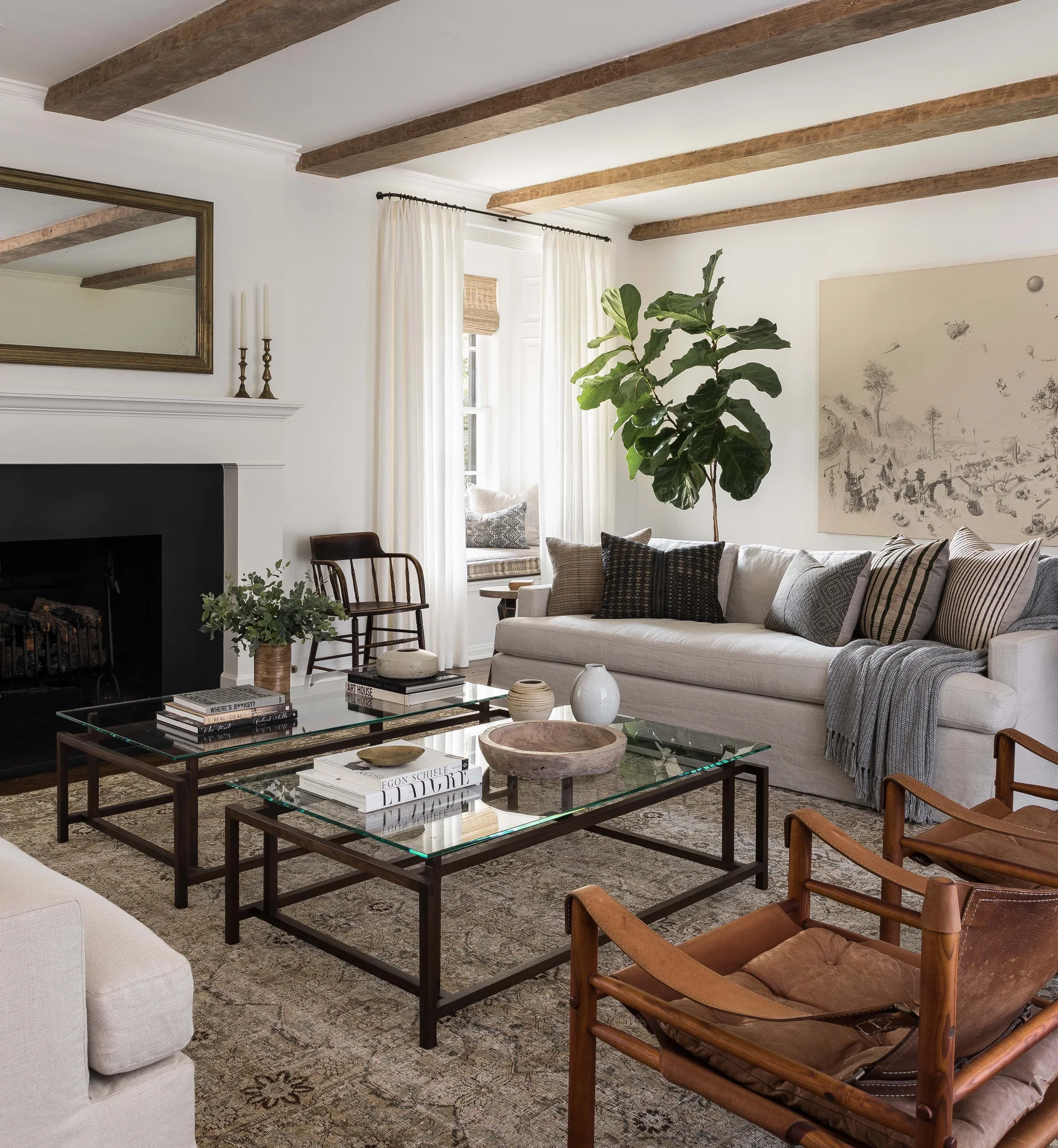 A bright living room features exposed wood ceiling beams, a cream sofa, two glass-topped coffee tables, and a fireplace with a black surround.