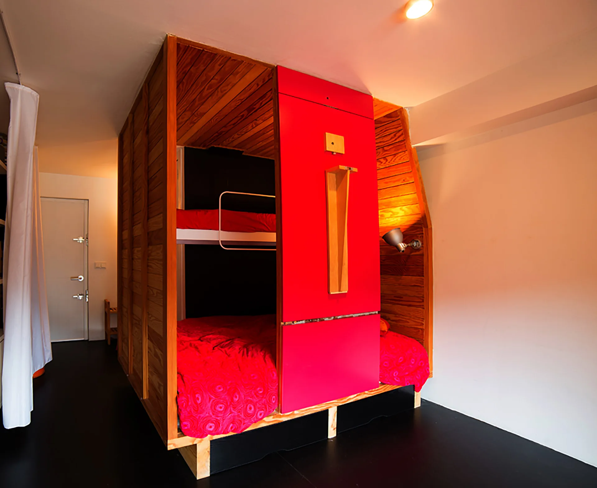 A custom built-in wooden bunk bed structure features warm wood paneling, a bright red front panel, and red bedding against white bedroom walls.