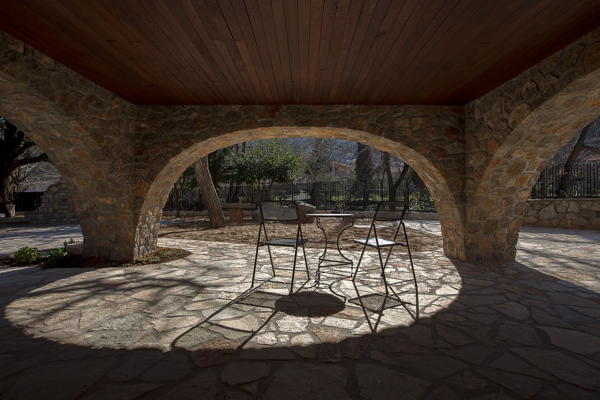 A shaded outdoor patio with stone archways, a dark wood ceiling, flagstone flooring, and a black metal bistro set looking out to a garden.