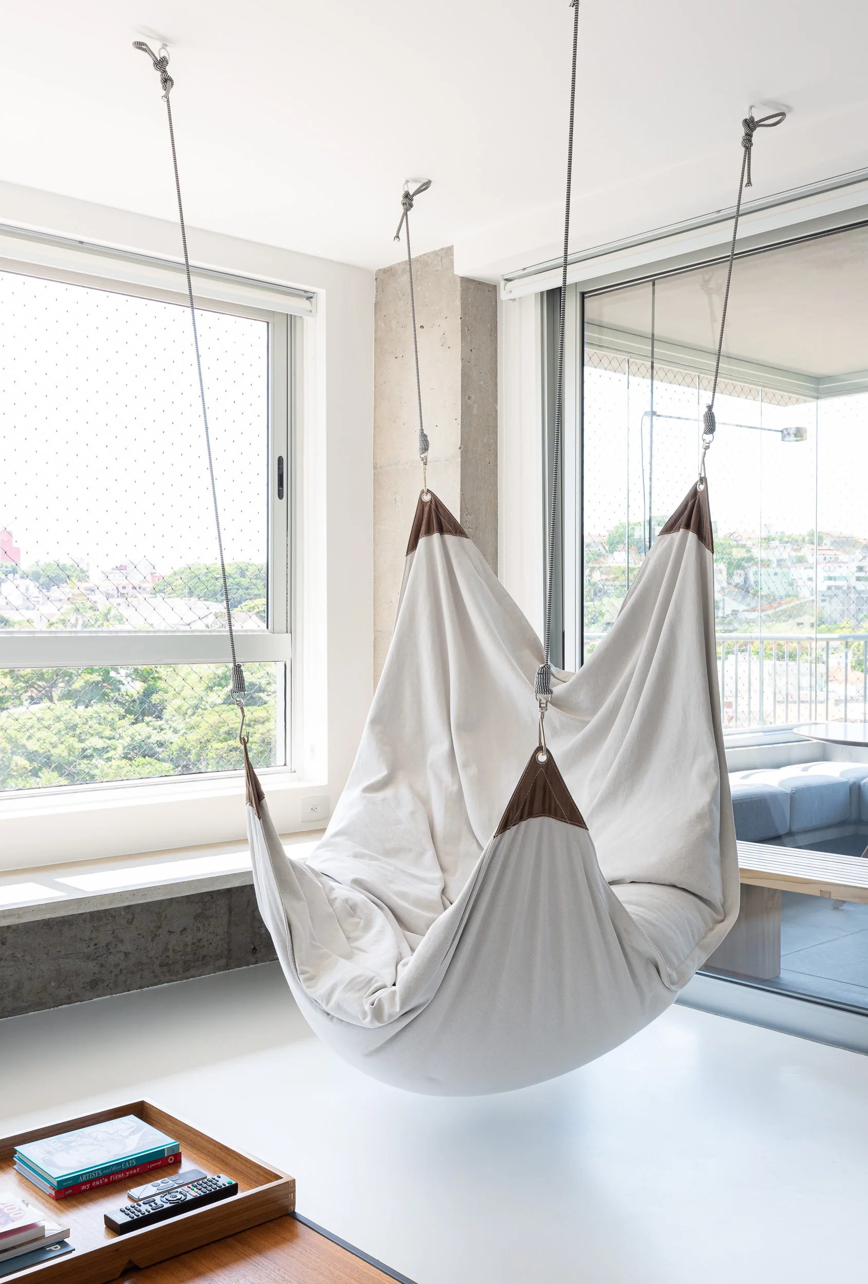 An off-white canvas hammock chair hangs from the ceiling in a minimalist white living room next to large windows and a raw concrete column.