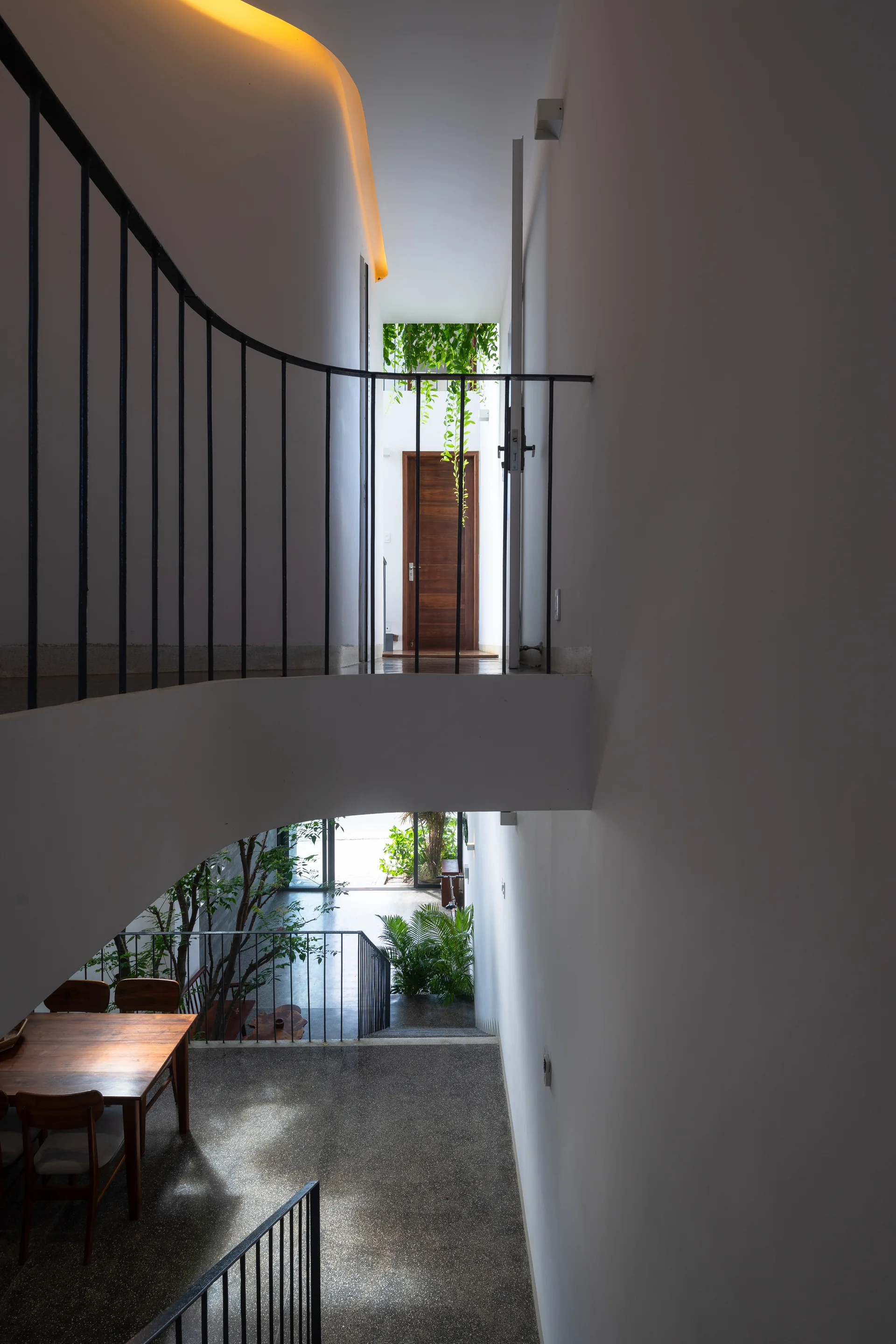 A view from a modern white upper landing with black metal railings looking down at a dining area, speckled terrazzo flooring, and indoor plants.