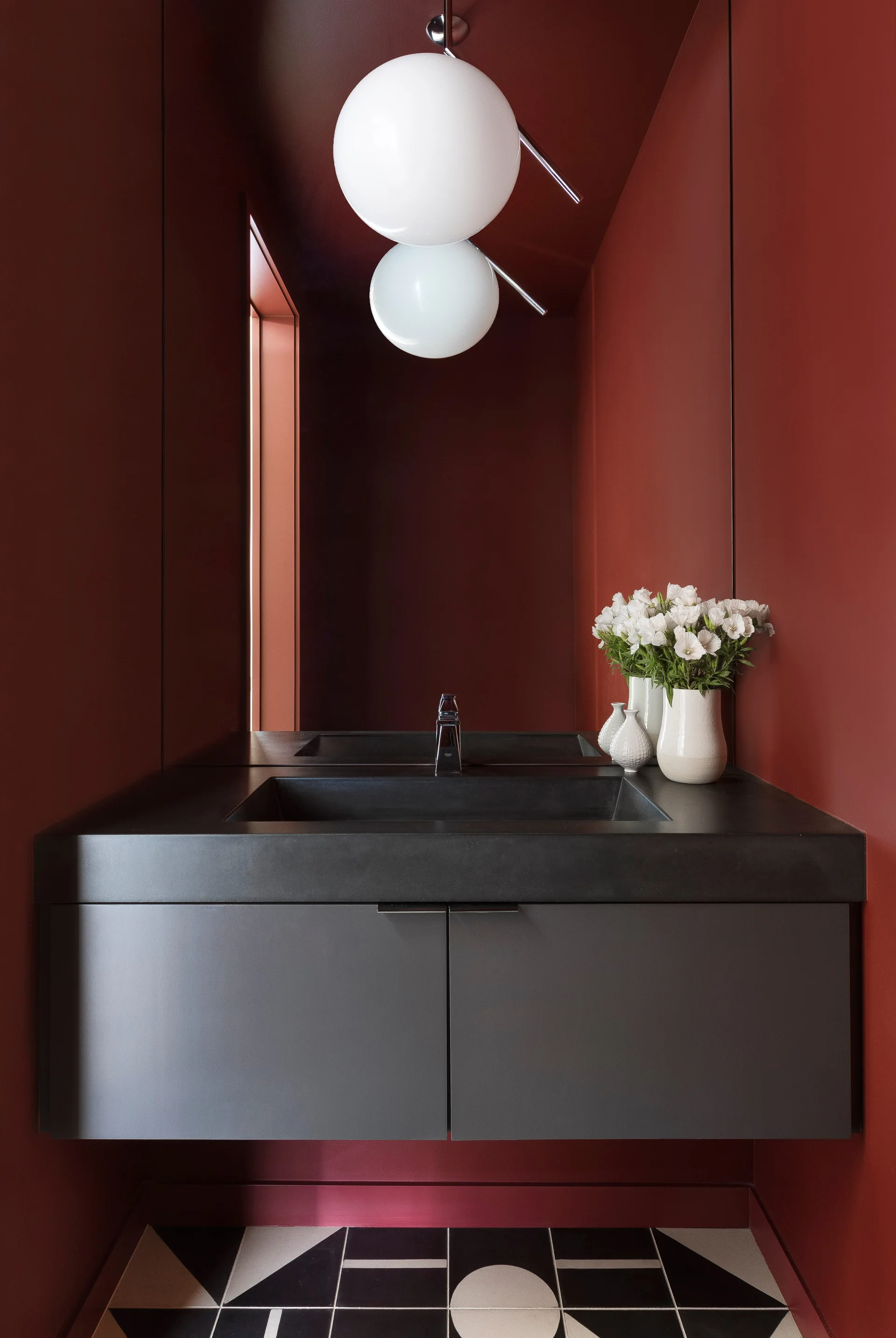 A modern powder room features deep red walls, a black floating vanity with an integrated sink, geometric floor tile, and a double globe pendant light.