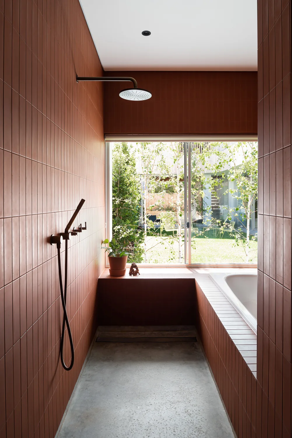 A modern wet room featuring vertical terracotta tiles on the walls, a raw concrete floor, and a large window looking out onto a green garden.