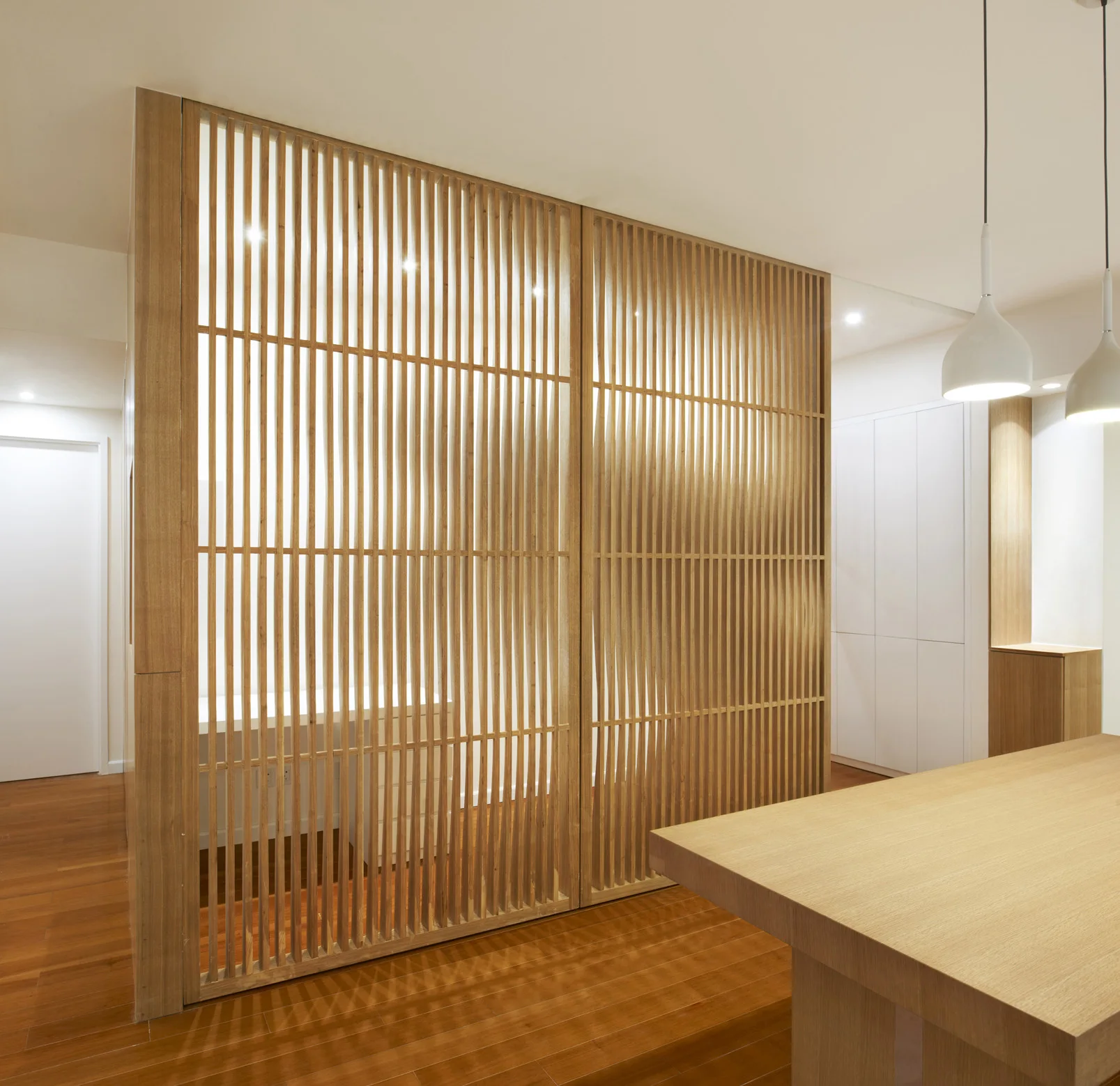 A modern interior featuring a floor-to-ceiling slatted wood partition next to a large wooden table under white pendant lights.