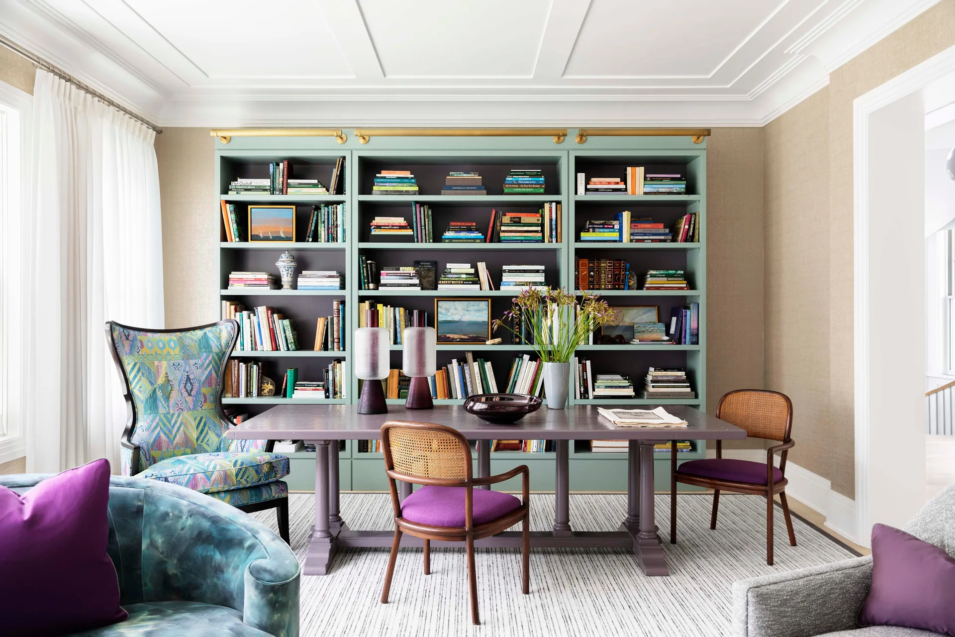 A home library featuring mint green built-in bookshelves, a purple wooden table with cane-back chairs, and textured beige wallpaper.