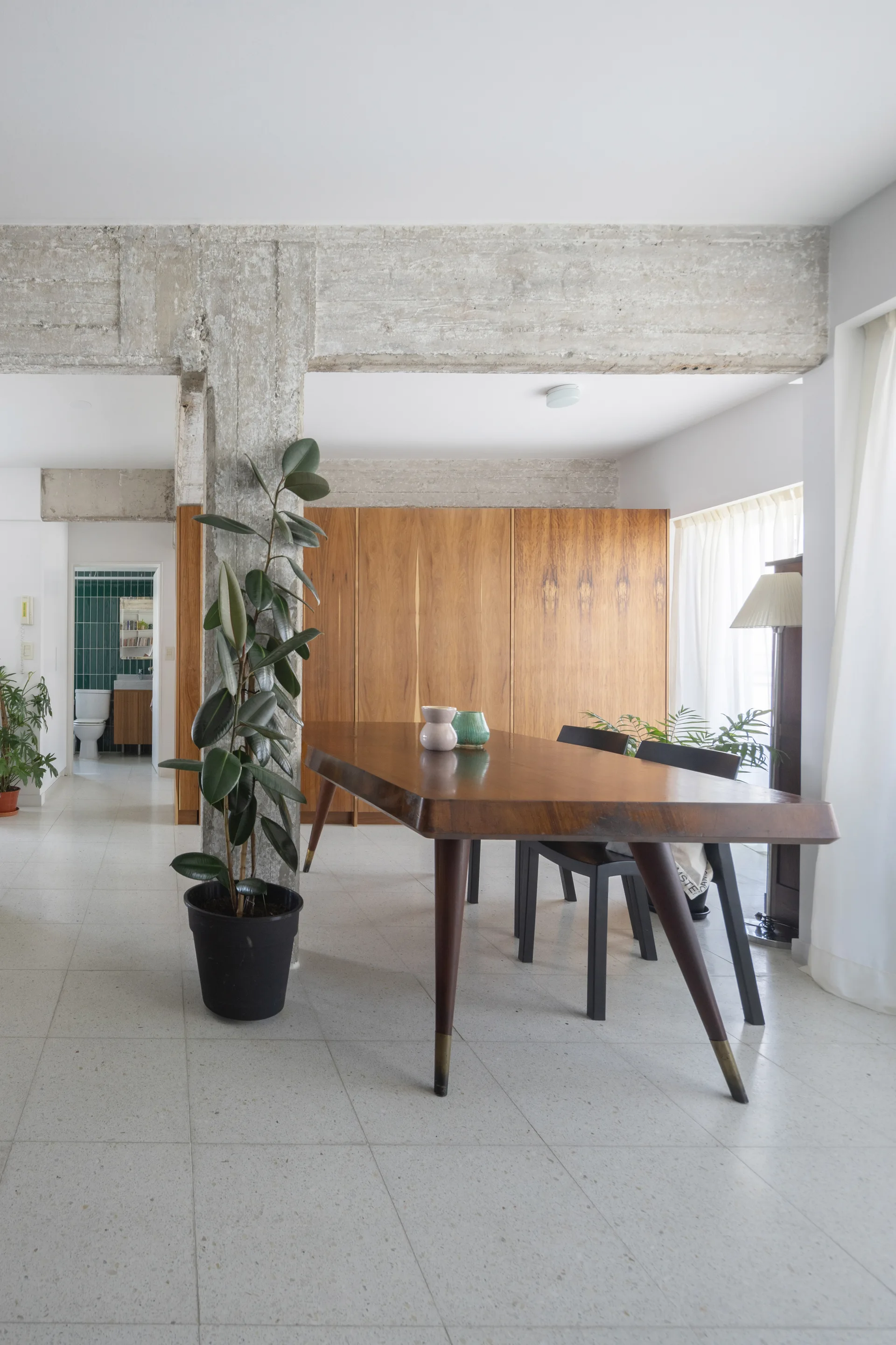 A minimalist dining room features a solid wood table with chairs under an exposed concrete beam next to a large potted plant and wood cabinets.