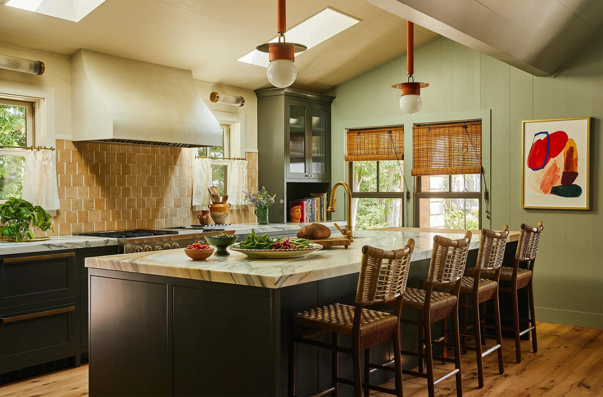 A kitchen features a dark gray island with a marble countertop, woven bar stools, sage green wood-paneled walls, and pendant lights.