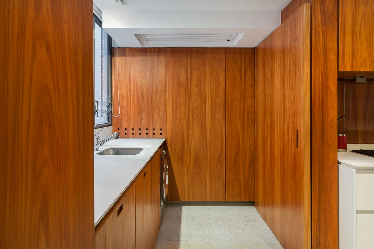 A laundry room features full-height warm wood-paneled storage, a white countertop with an undermount sink, and light speckled floor tile.