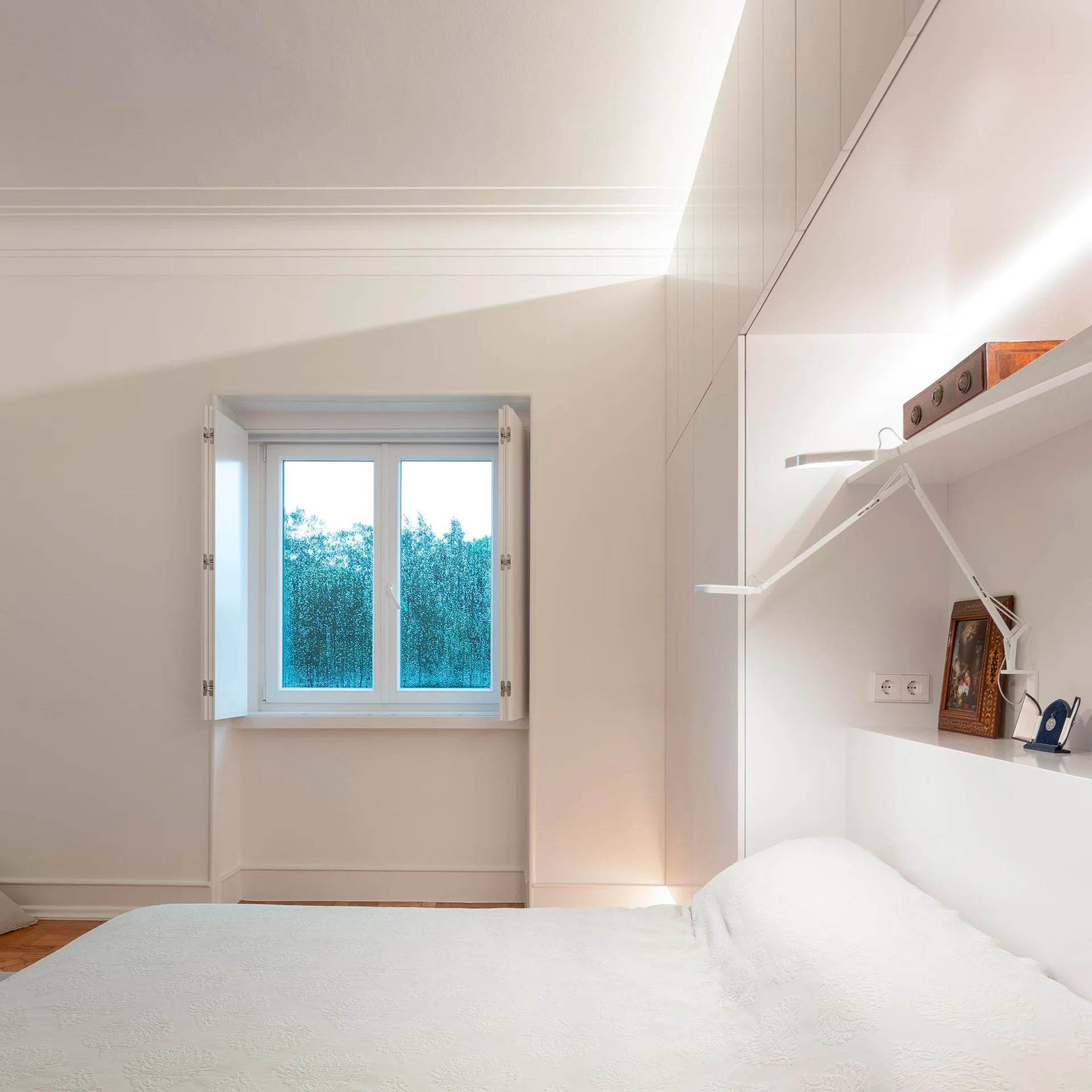 A minimalist white bedroom features a bed tucked under built-in glossy storage cabinets, integrated lighting, and a window with open shutters.