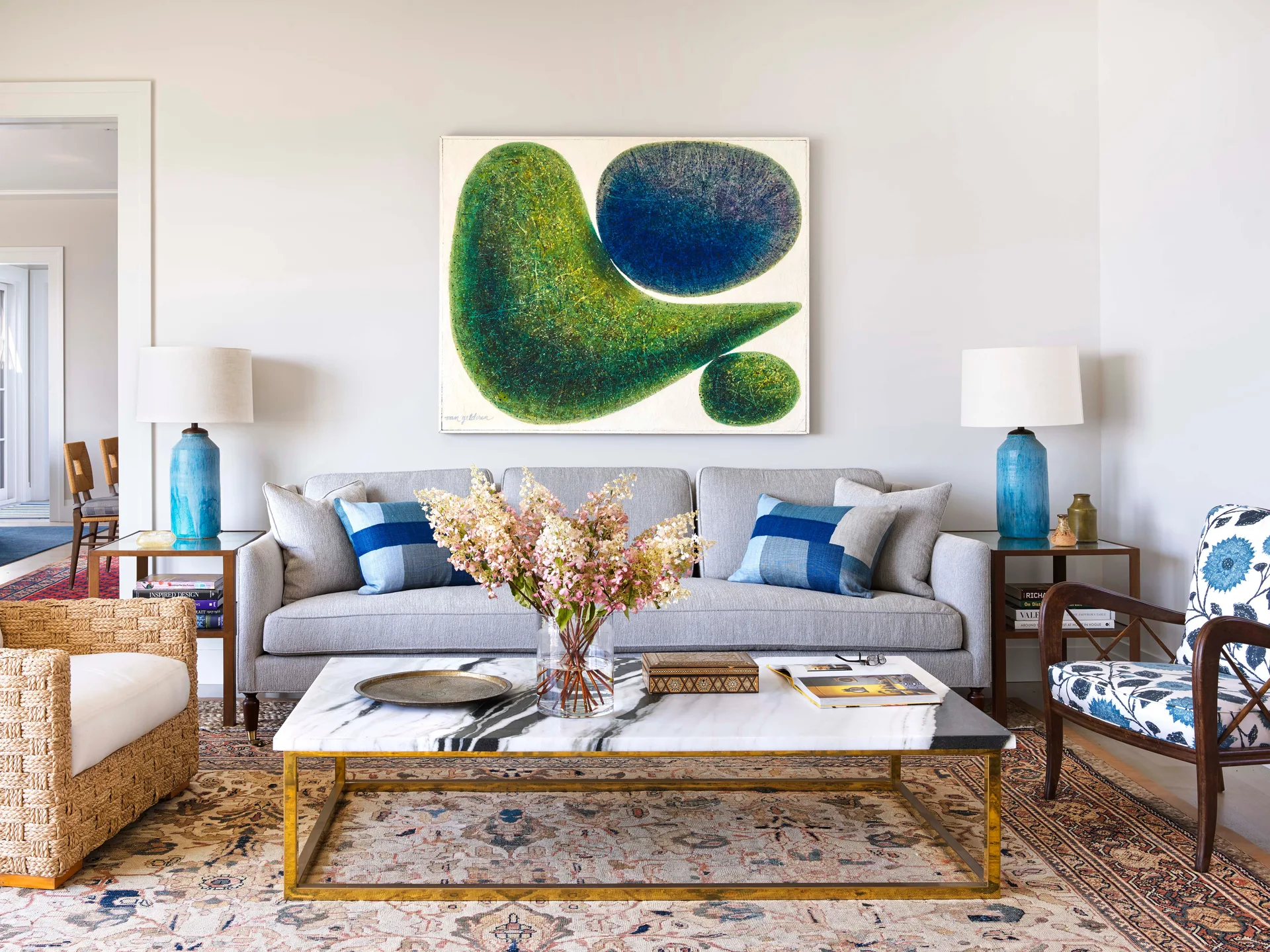 A grey sofa sits below a green abstract painting, flanked by blue table lamps and set before a marble coffee table on a traditional rug.