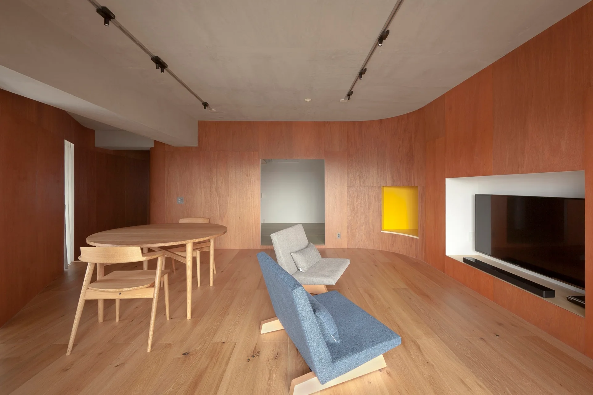 A minimalist living room features wood-paneled walls, a round dining table, two low lounge chairs, and a built-in TV niche.