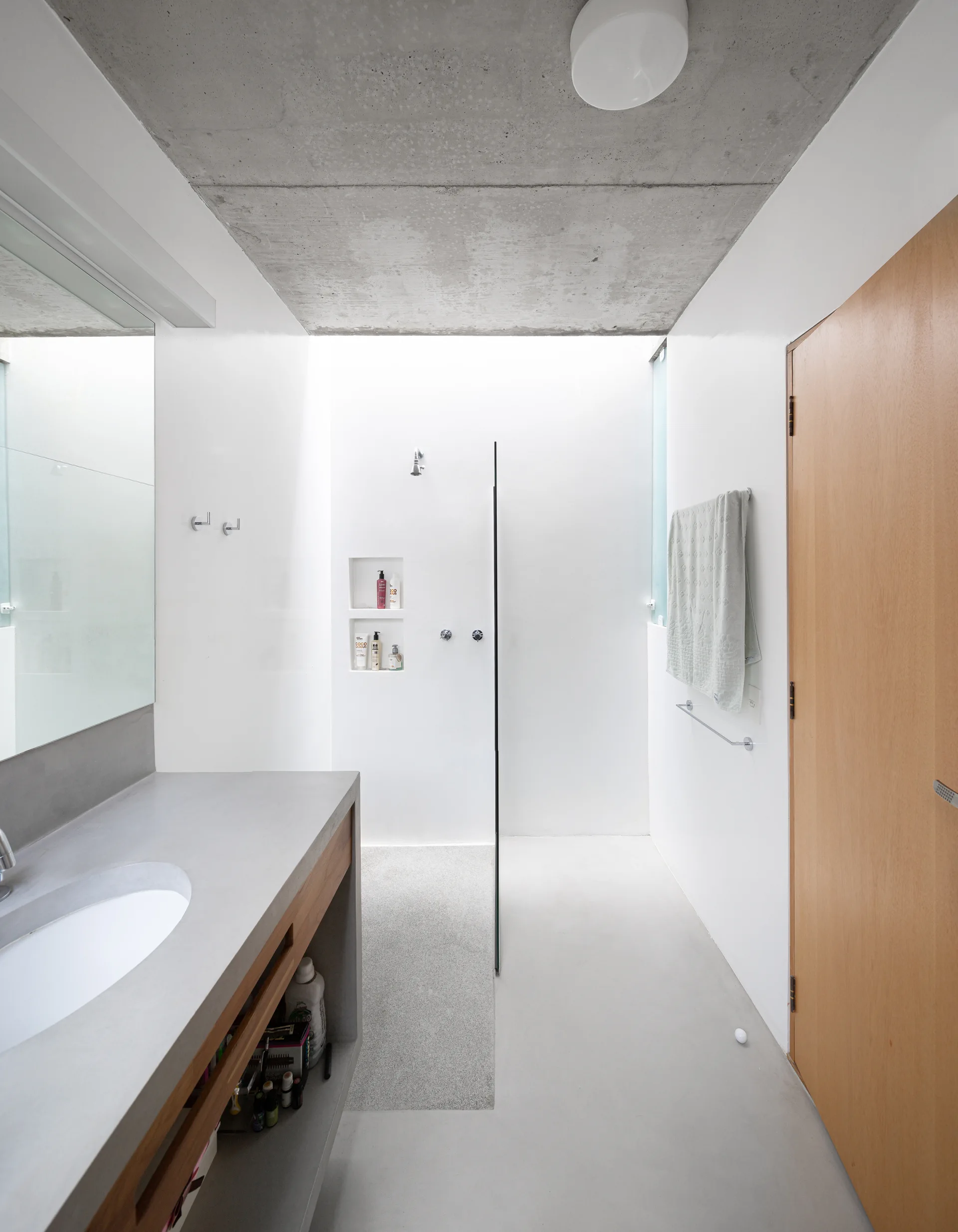 A minimalist bathroom features a textured concrete ceiling, a custom concrete vanity with an integrated sink, white walls, and a glass-walled shower.