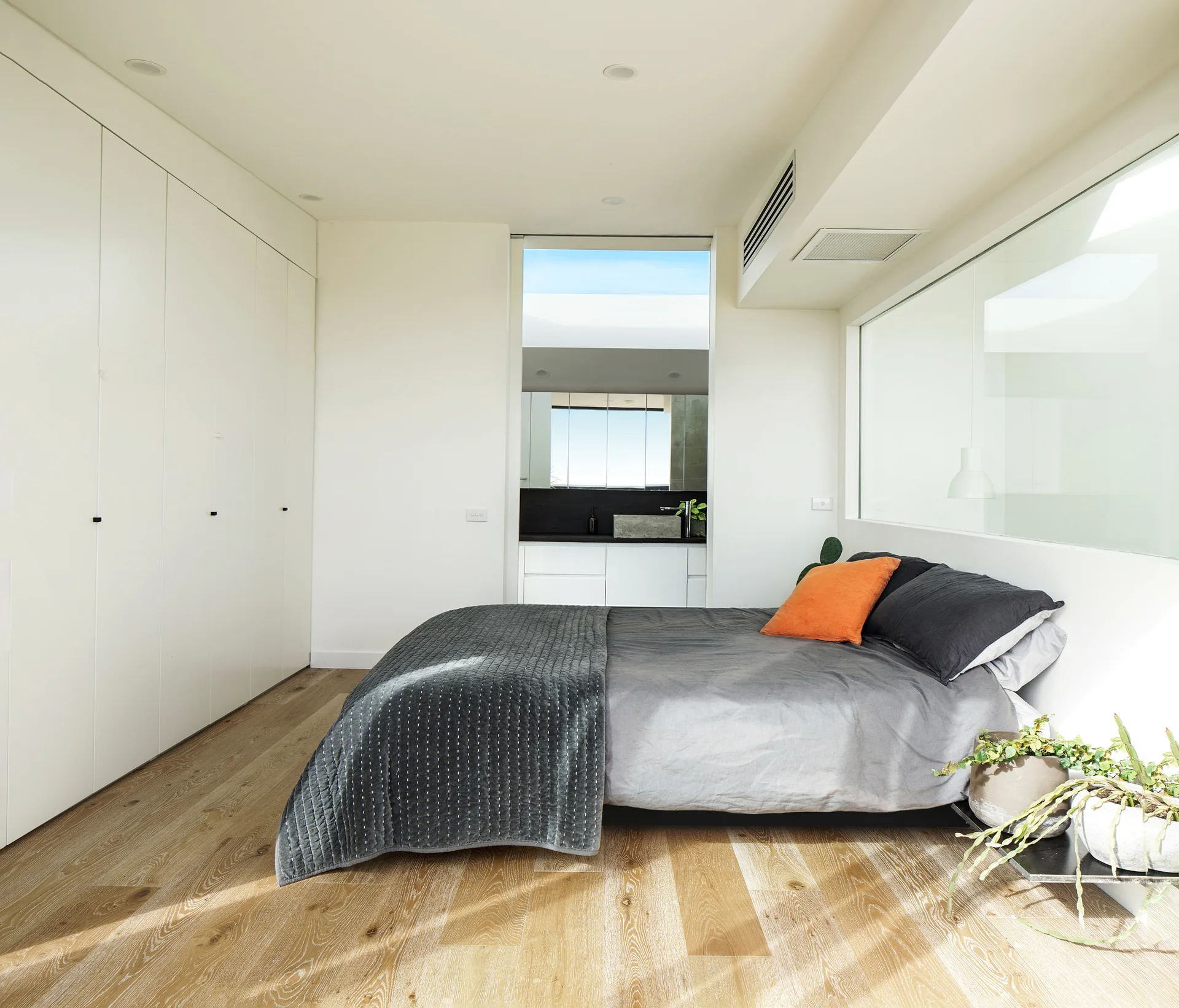 A minimalist bedroom features a bed with grey bedding and an orange pillow, next to a wall of white built-in closets and light wood floors.