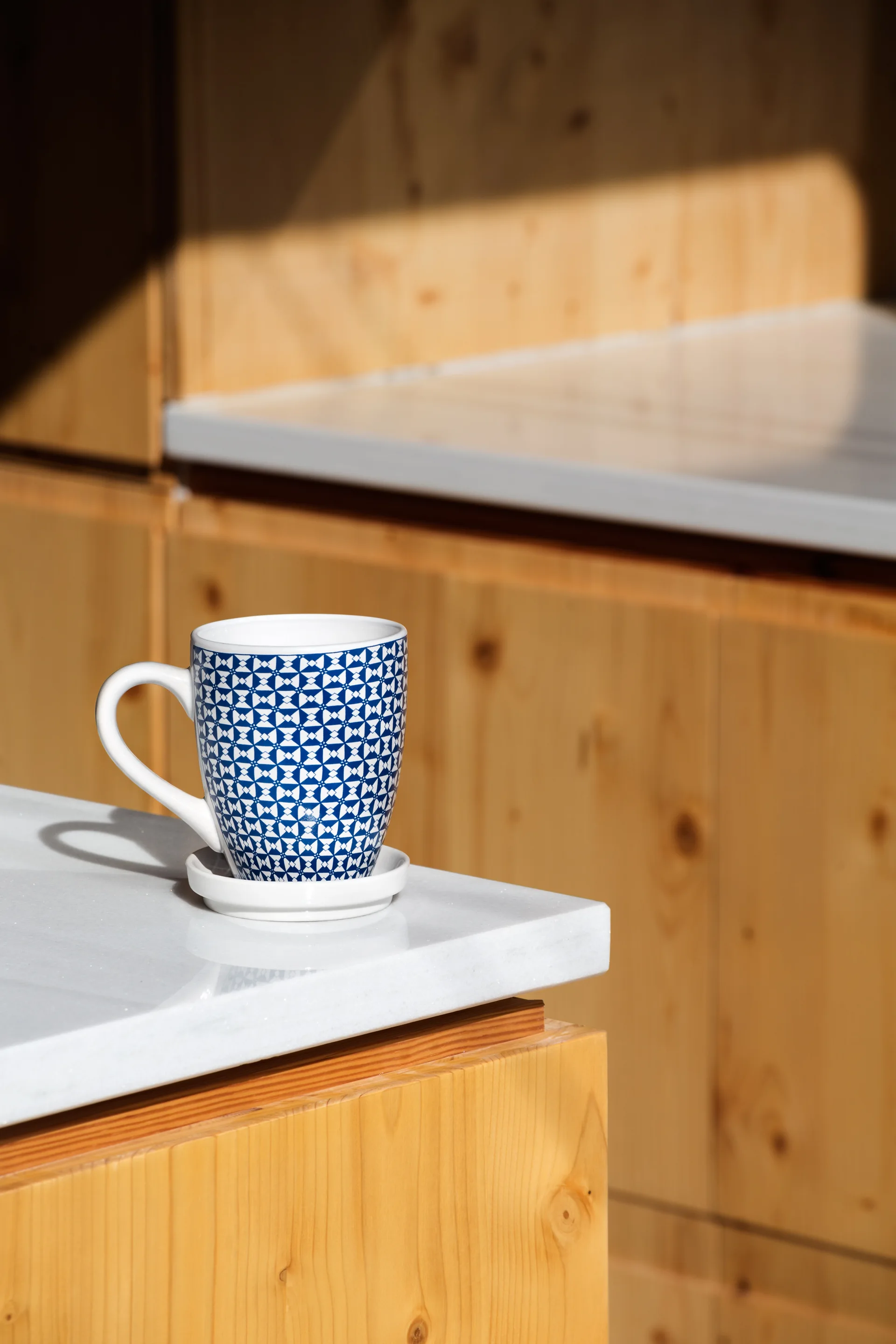 A patterned blue and white ceramic mug rests on a white saucer atop a stone countertop with light pine wood cabinets in the background.