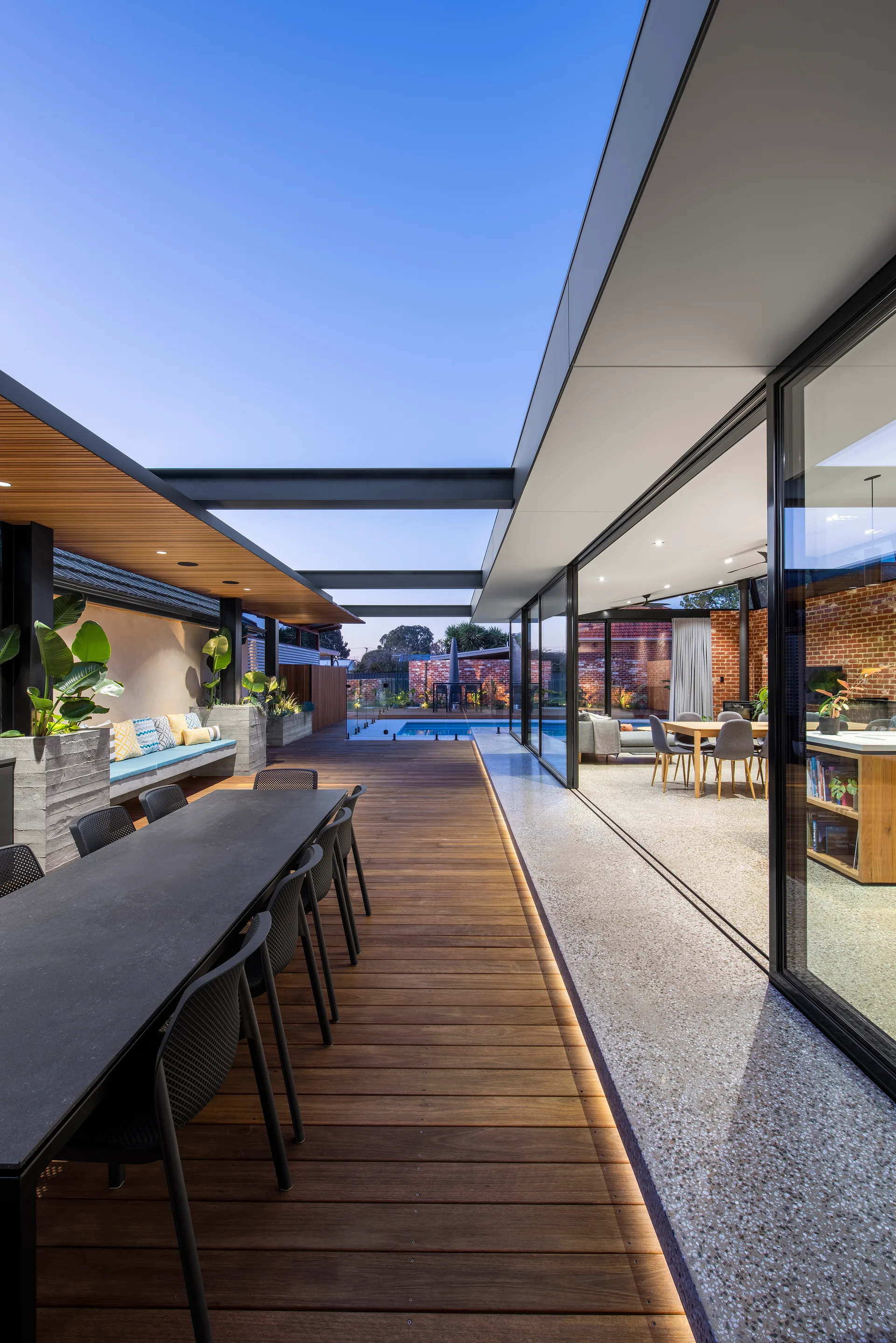 A modern outdoor timber deck features a long dining table and chairs next to sliding glass doors that open to an indoor dining area.