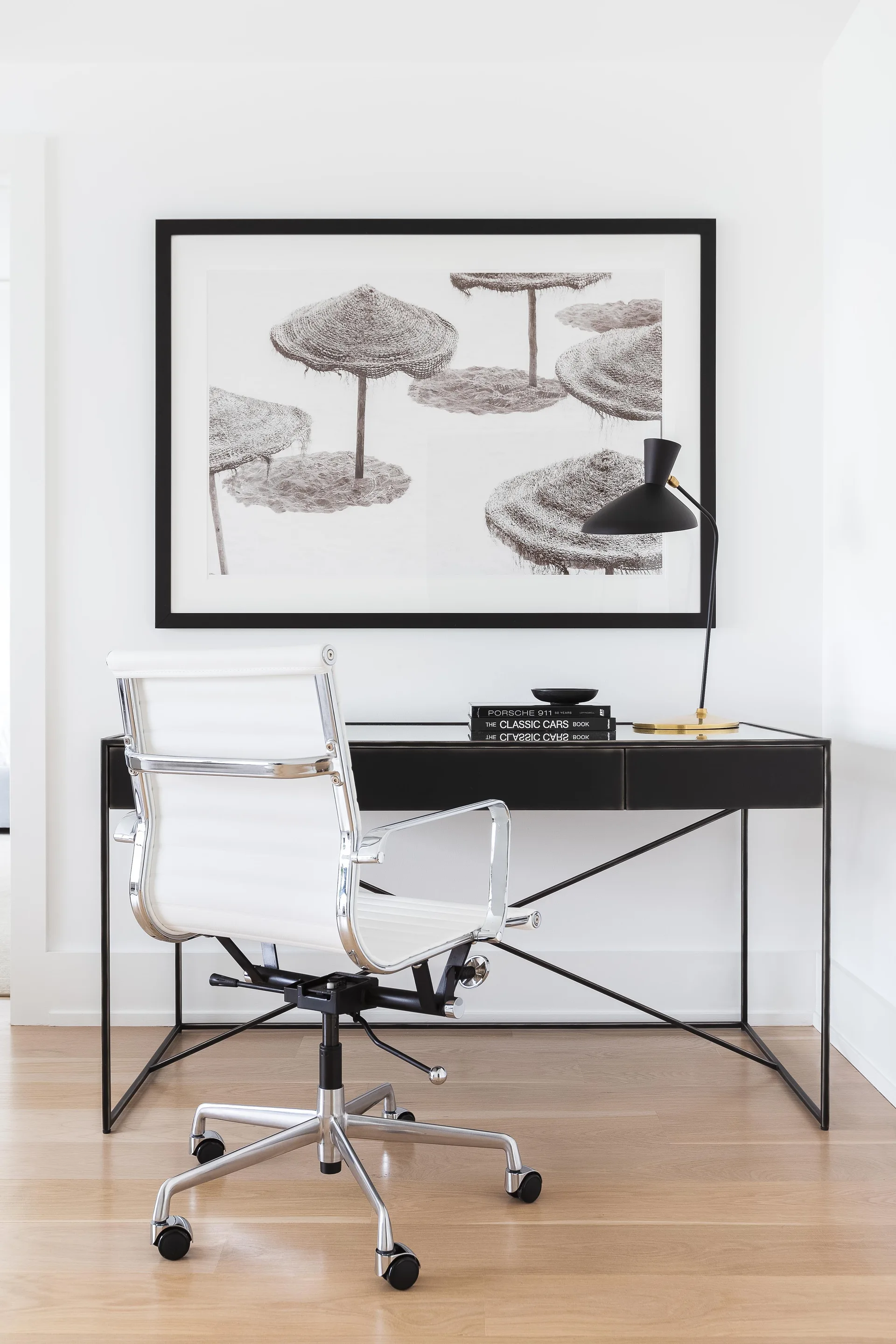 A modern home office features a white leather office chair pulled up to a black metal desk beneath large framed black-and-white art.