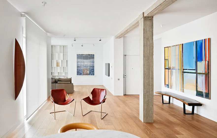 A minimalist living room featuring an exposed concrete column, light wood floors, two red leather chairs, and a large abstract wall painting.