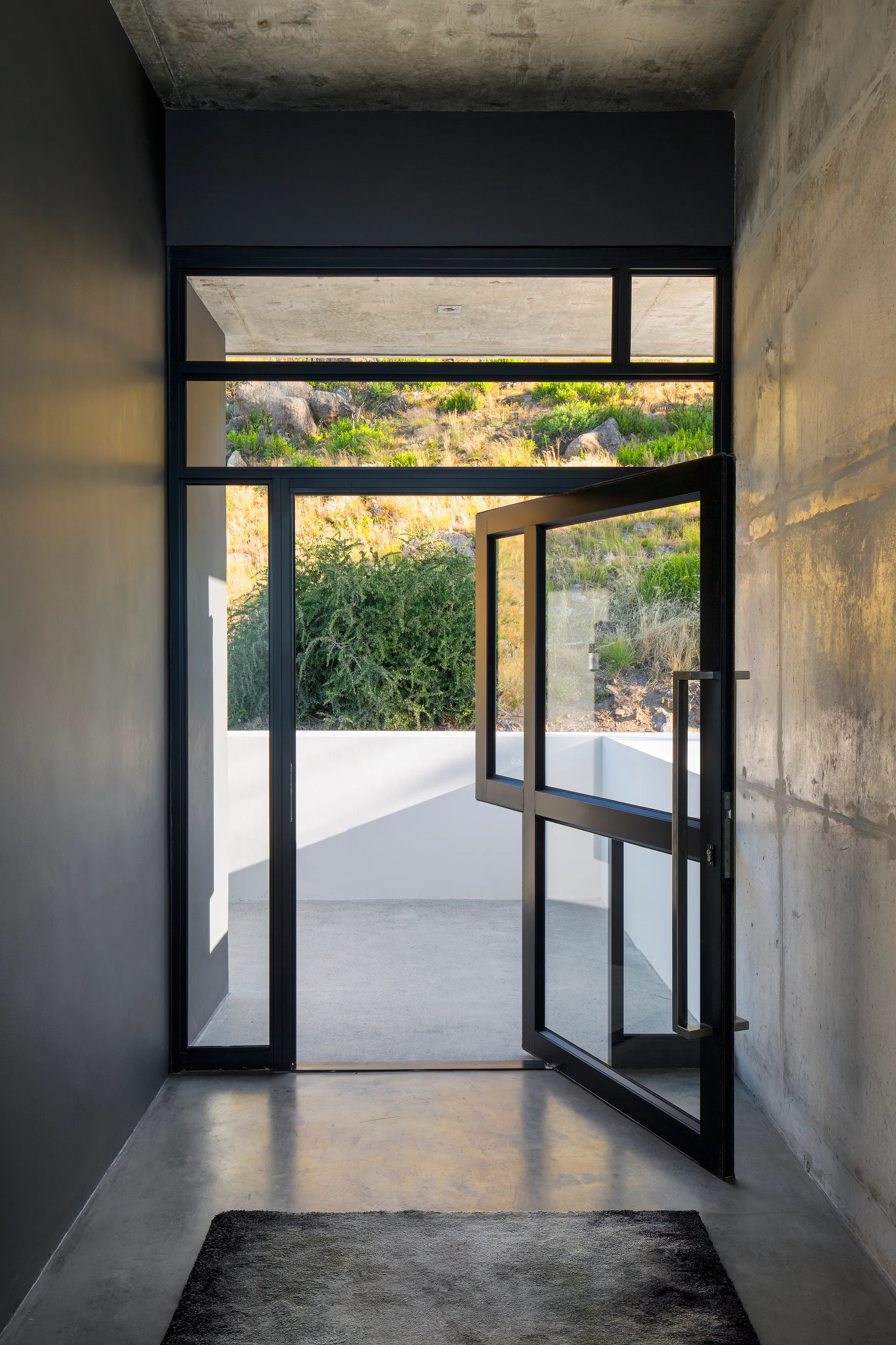 A minimalist entryway features a black-framed glass door, polished concrete floors, a raw concrete wall, and a dark grey accent wall.