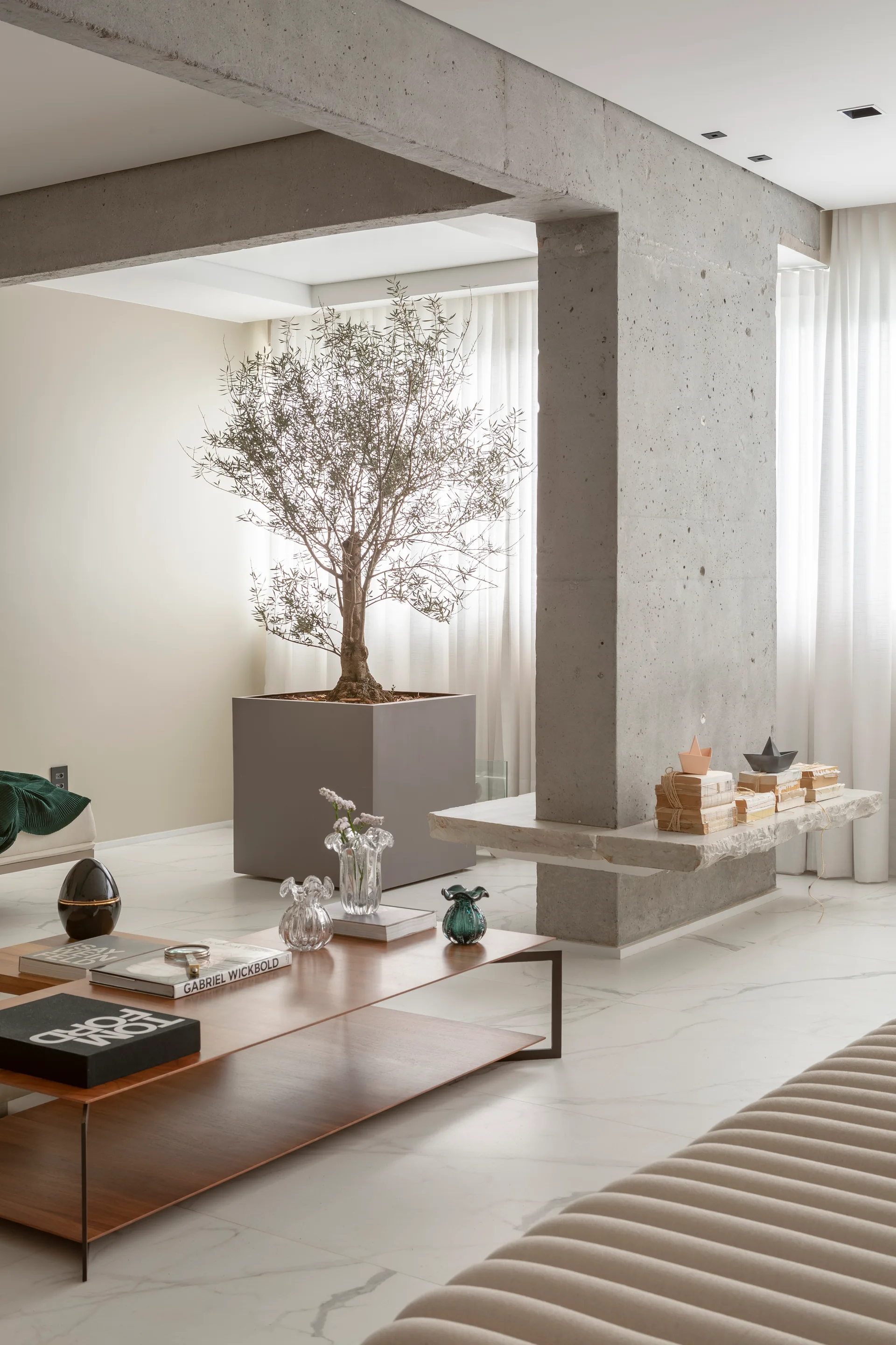 A minimalist living room features an exposed concrete pillar with a stone ledge, a potted olive tree, and a wooden coffee table.