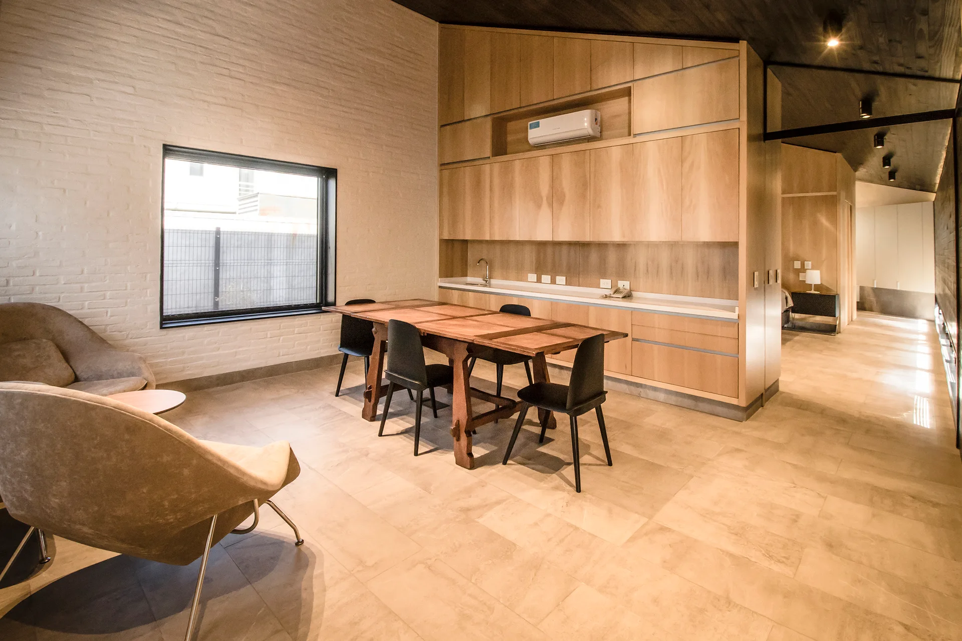 A dining room features a wooden table and black chairs beside a white painted brick wall and a large light wood cabinetry unit.