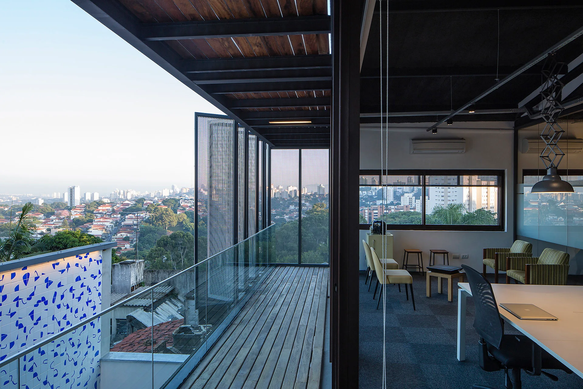 A modern office room opens onto a wooden balcony featuring a glass railing and tall perforated metal screens overlooking a distant city skyline.